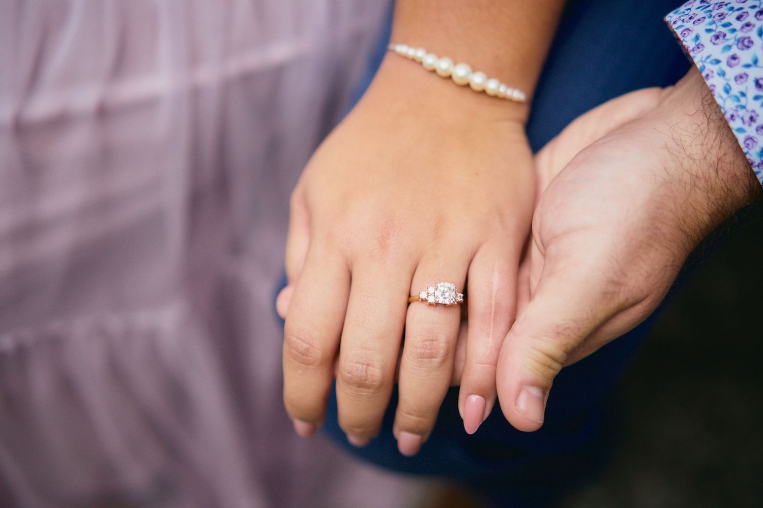 Close up view of hands showing an engagement ring and pearl bracelet against a soft purple background.