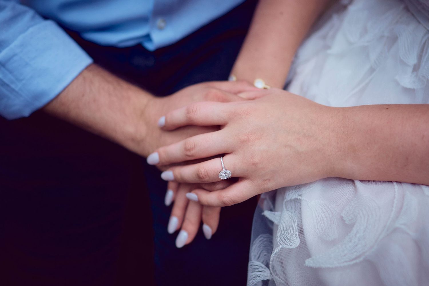 Close-up of two hands wearing wedding rings with white lace dress and blue suit fabric visible in background.