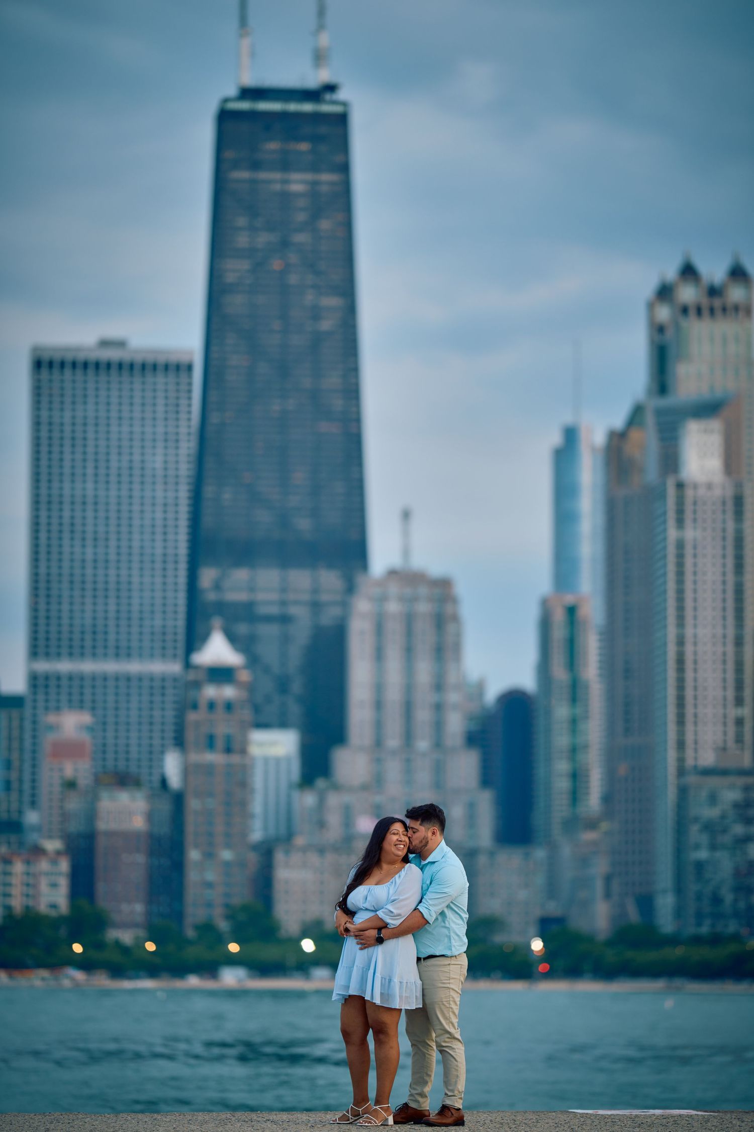 Couple embraces on North Avenue Beach with the iconic John Hancock Center and Chicago skyline in background at dusk.