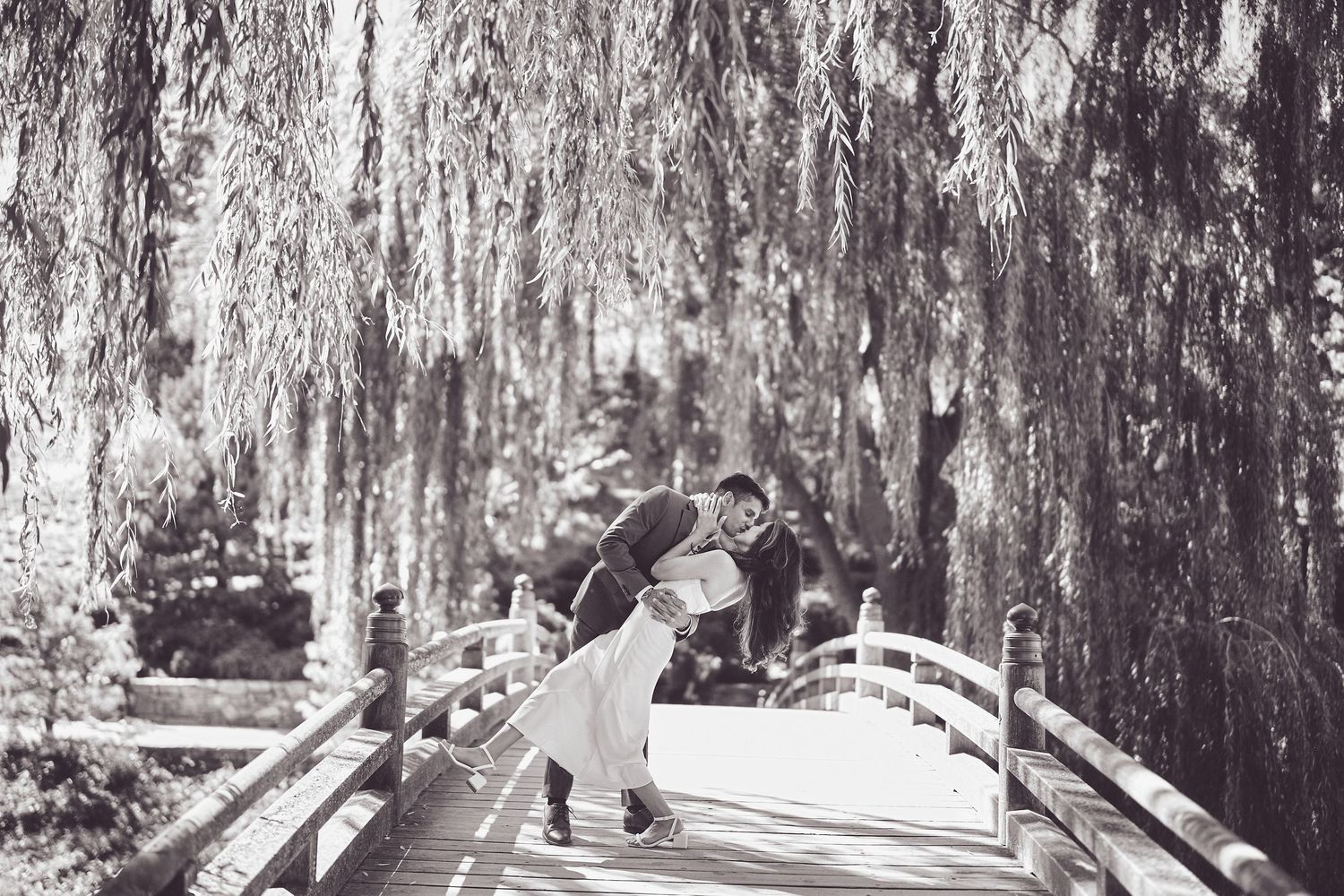 A couple shares a romantic moment on a white bridge under dramatic willow trees in black and white.