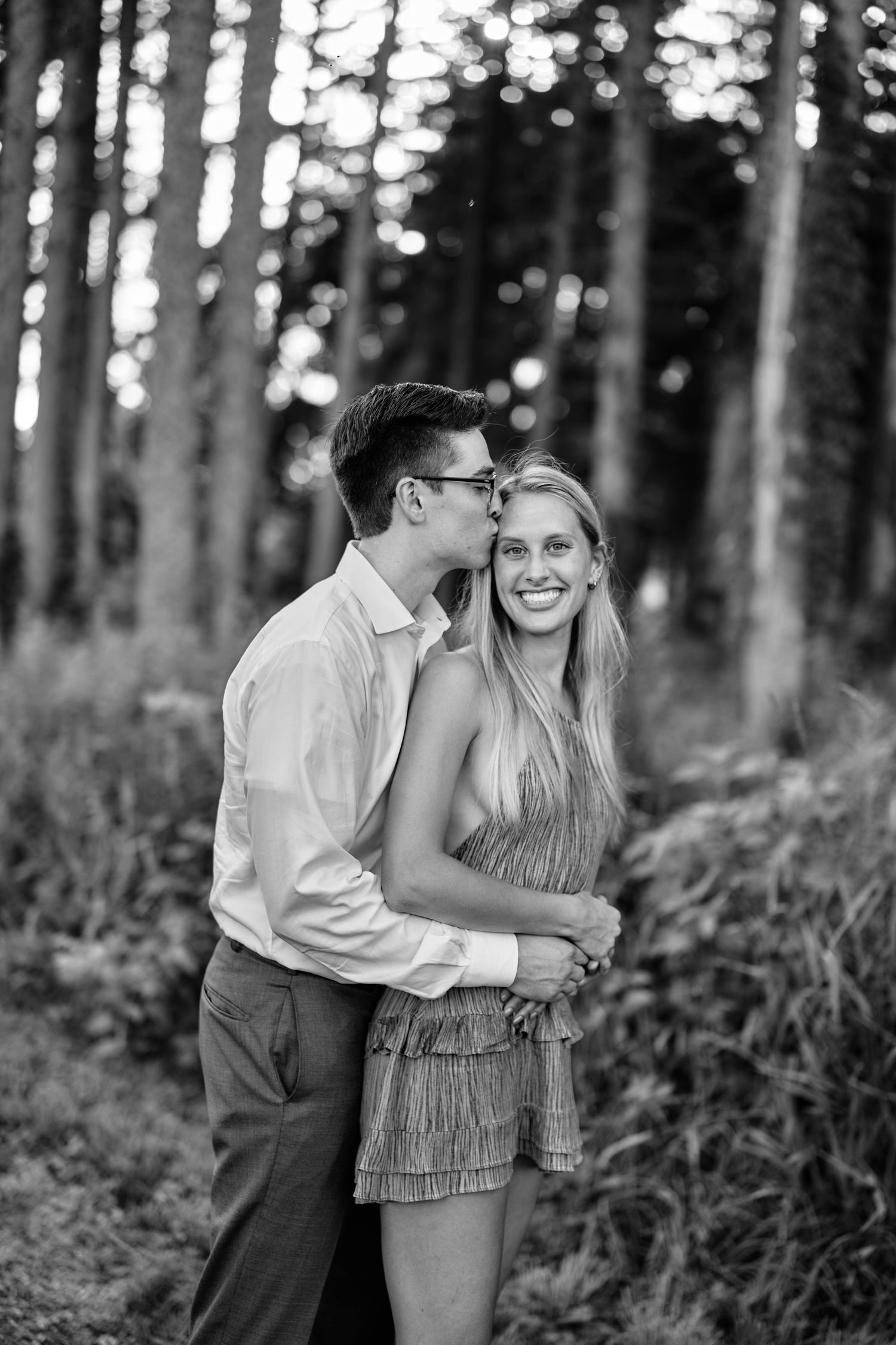 A romantic black and white forest portrait of a couple embracing among tall trees in an intimate outdoor setting.
