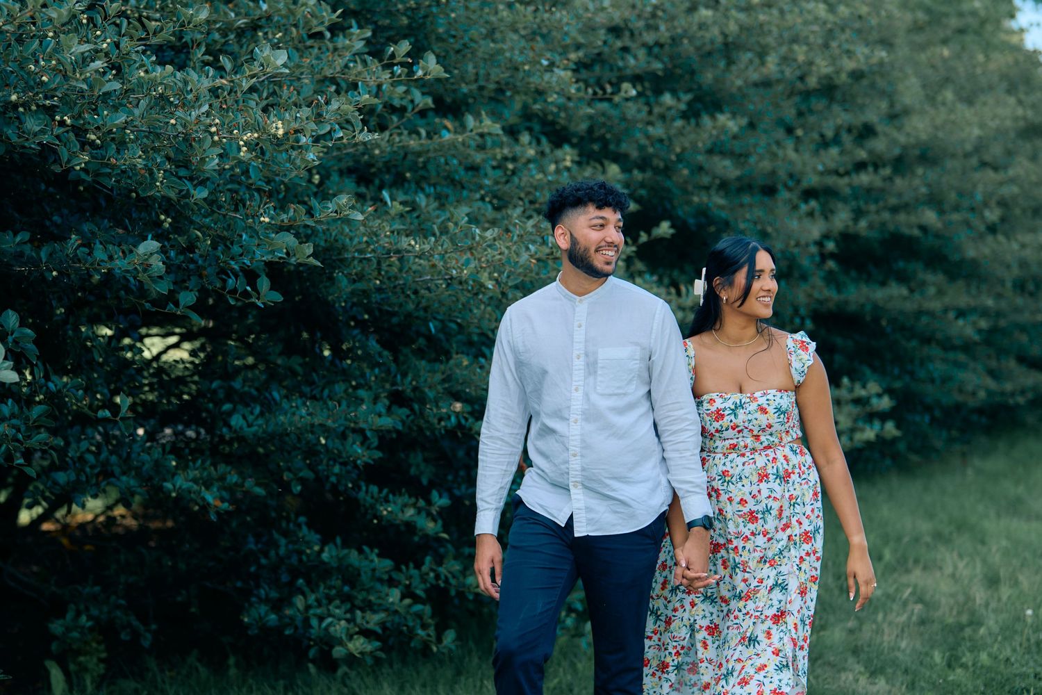 A couple walking together outdoors, with her in a floral dress and him in a light blue button-down shirt and hat.