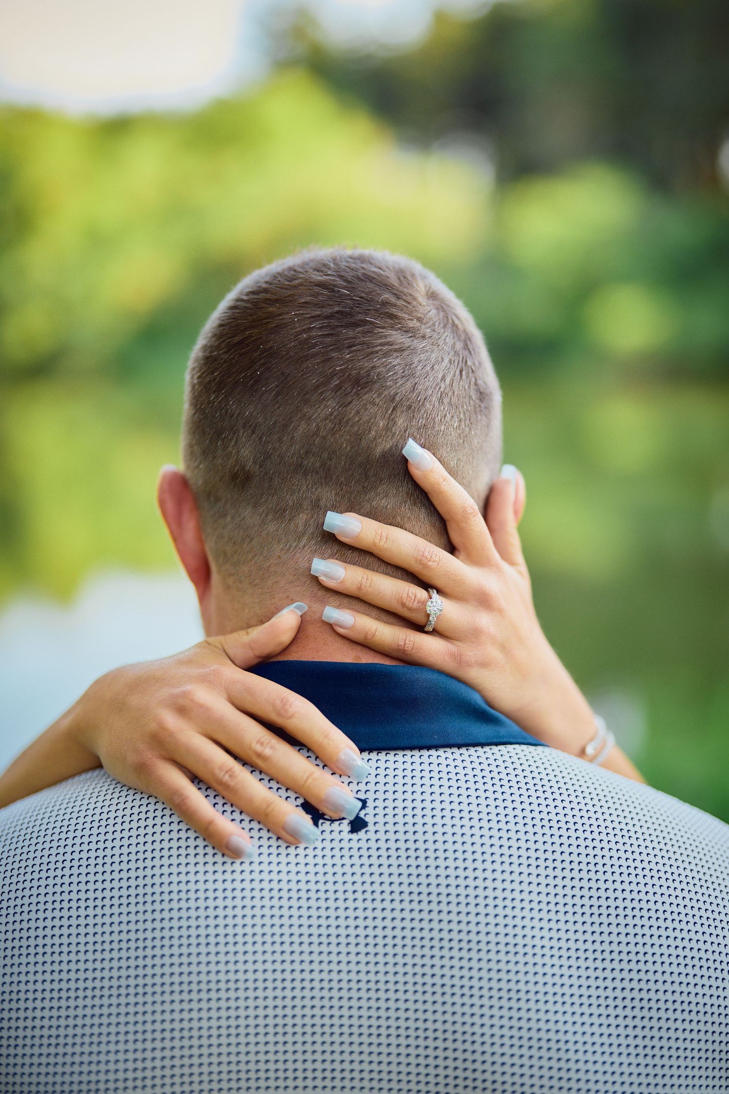 Hands with white nail polish and engagement ring embrace the back of someone wearing a gray patterned shirt outdoors.