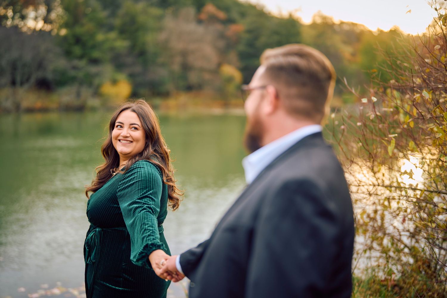 Romantic couple holds hands by a serene lake at sunset with fall foliage in the background.