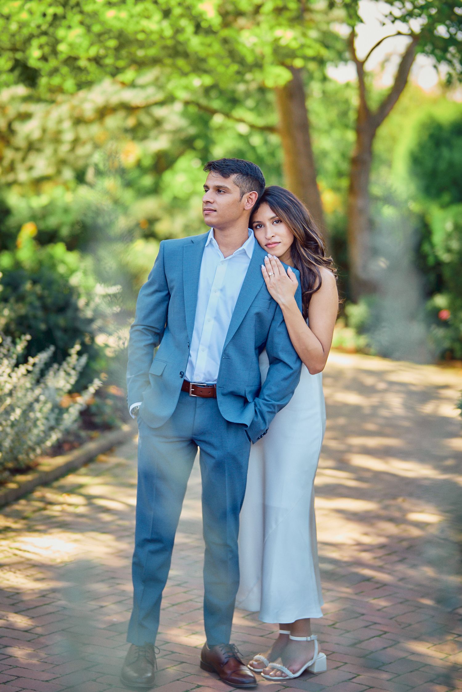 A couple poses together outdoors, with one wearing a blue suit and the other in a flowing light dress on a garden path.