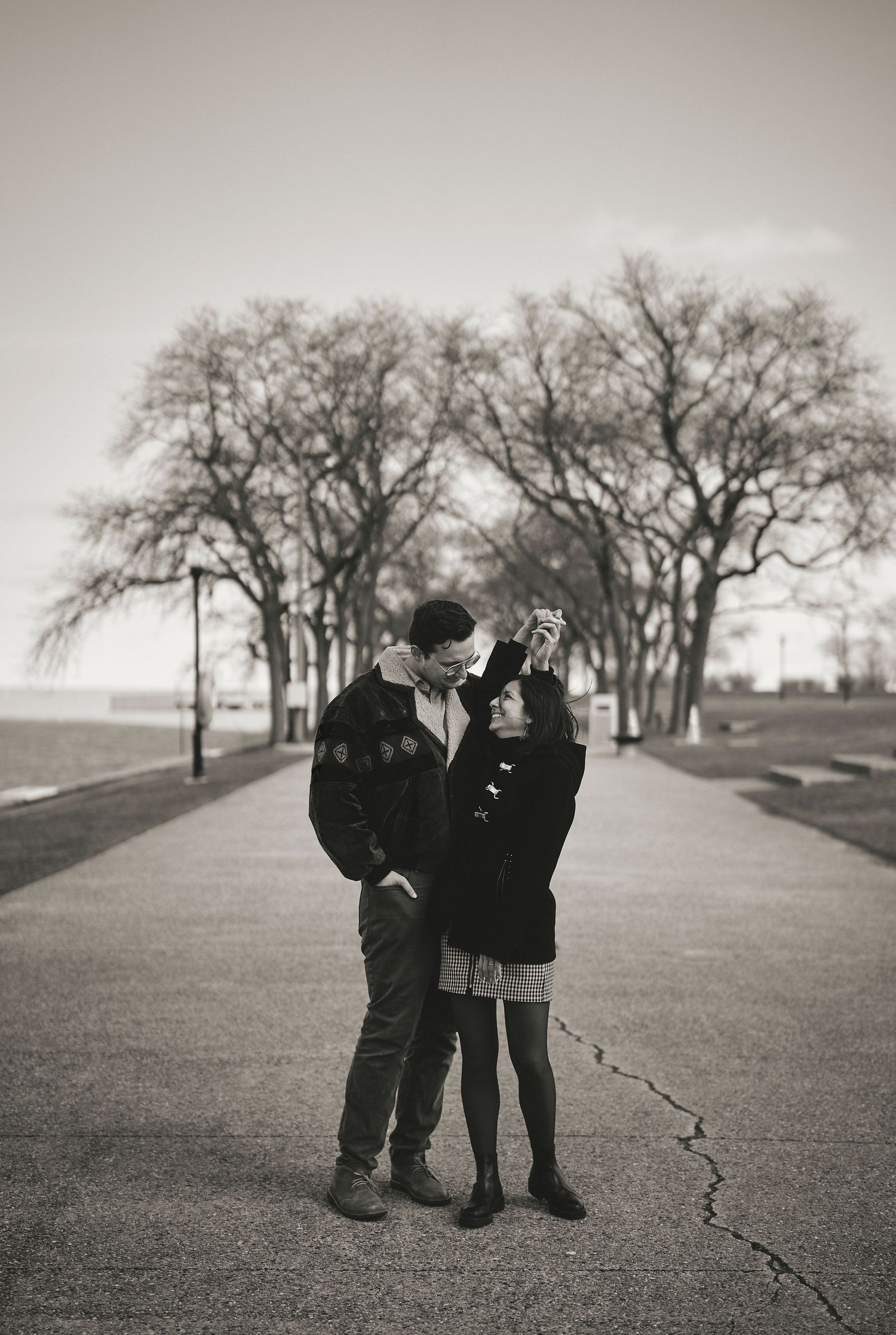 A couple shares an intimate moment while standing on a tree-lined pathway in black and white photography.