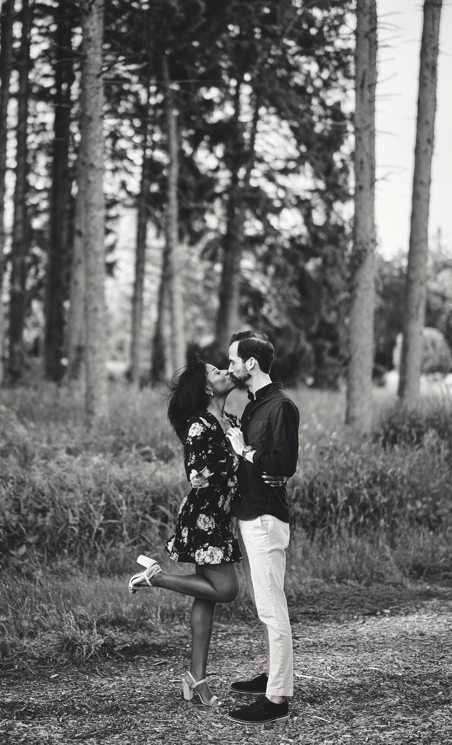 A romantic black and white photograph of a couple embracing in a forest setting, surrounded by tall trees.