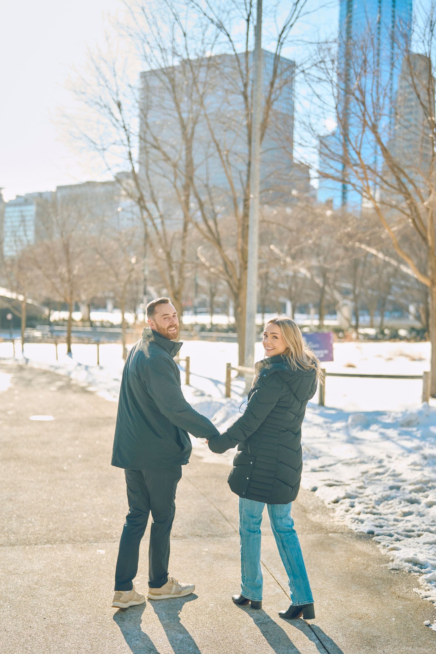 A couple holding hands walks along a snowy winter path in an urban park with city buildings visible in the background.