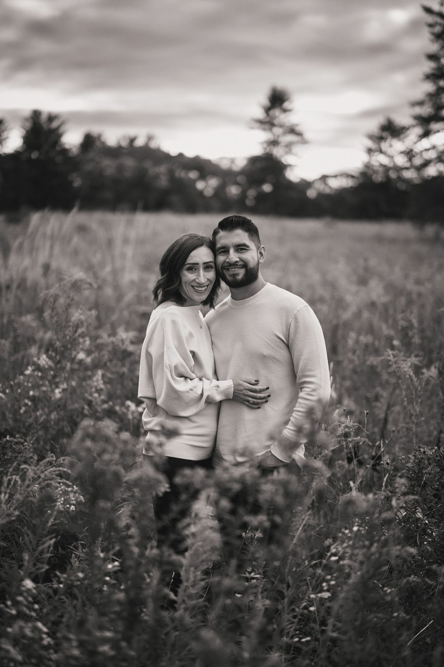 A couple stands together in a scenic field of wildflowers with trees and mountains in the background, shot in black and white.