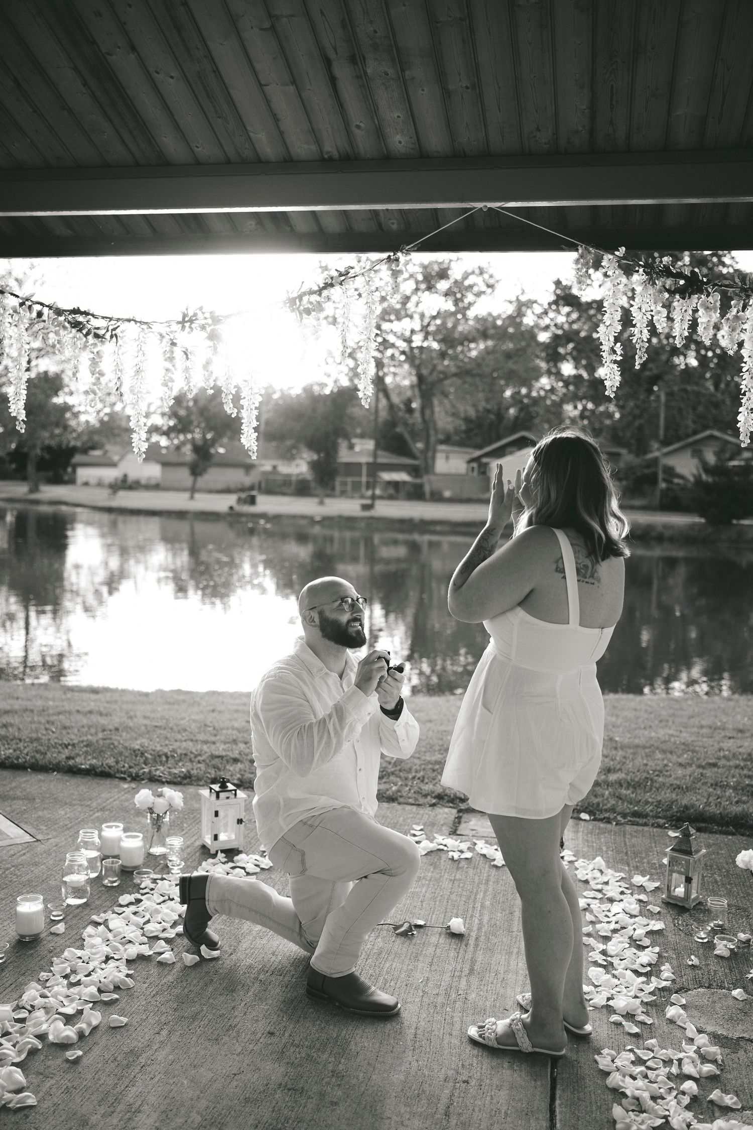 A romantic proposal scene on a lakeside deck decorated with candles and rose petals at sunset.