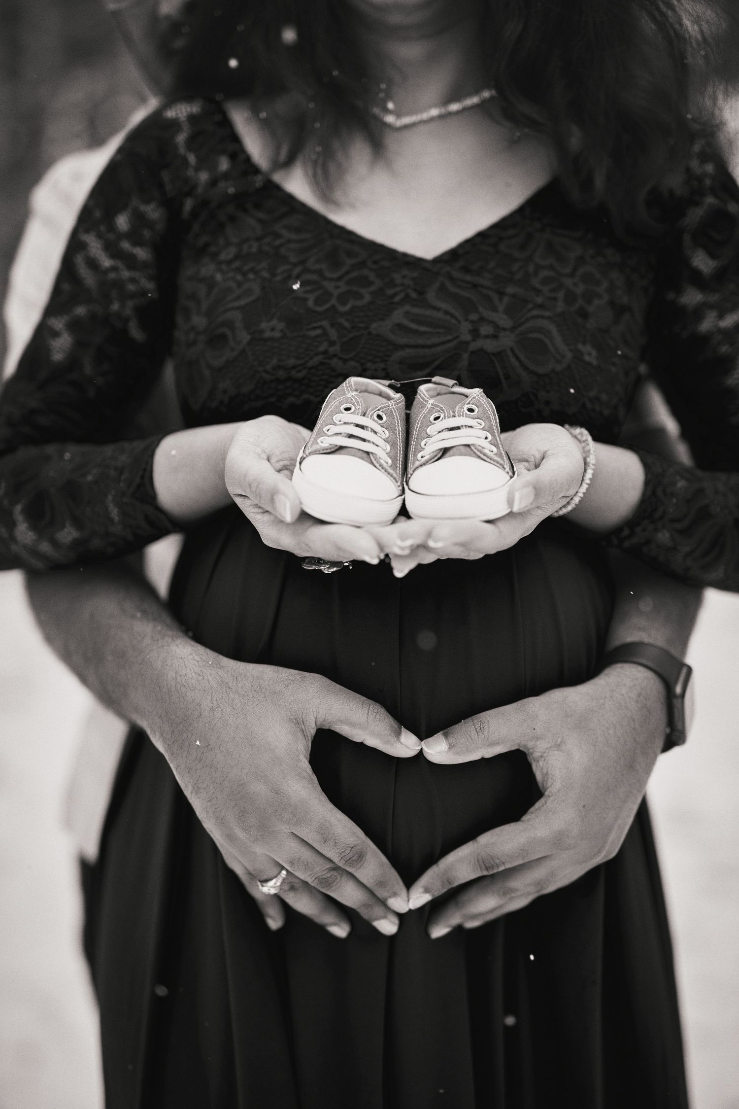 Black and white maternity photo showing hands holding tiny baby shoes while forming heart shape over pregnant belly.