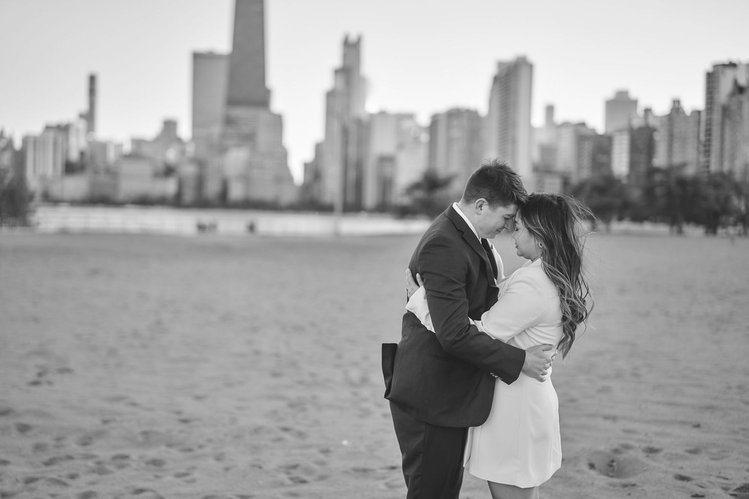 A romantic couple embraces on a sandy beach with the Chicago skyline visible across the water in black and white.