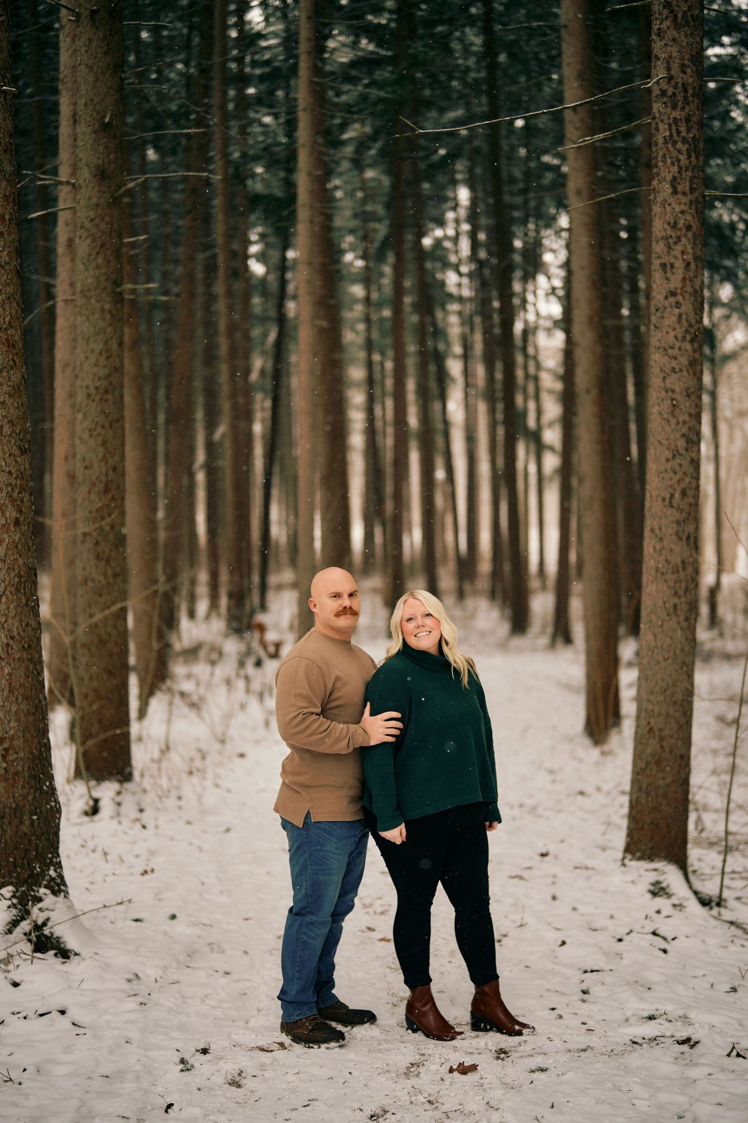 A couple stands embracing on a snowy path through a dense winter forest in warm sweaters.