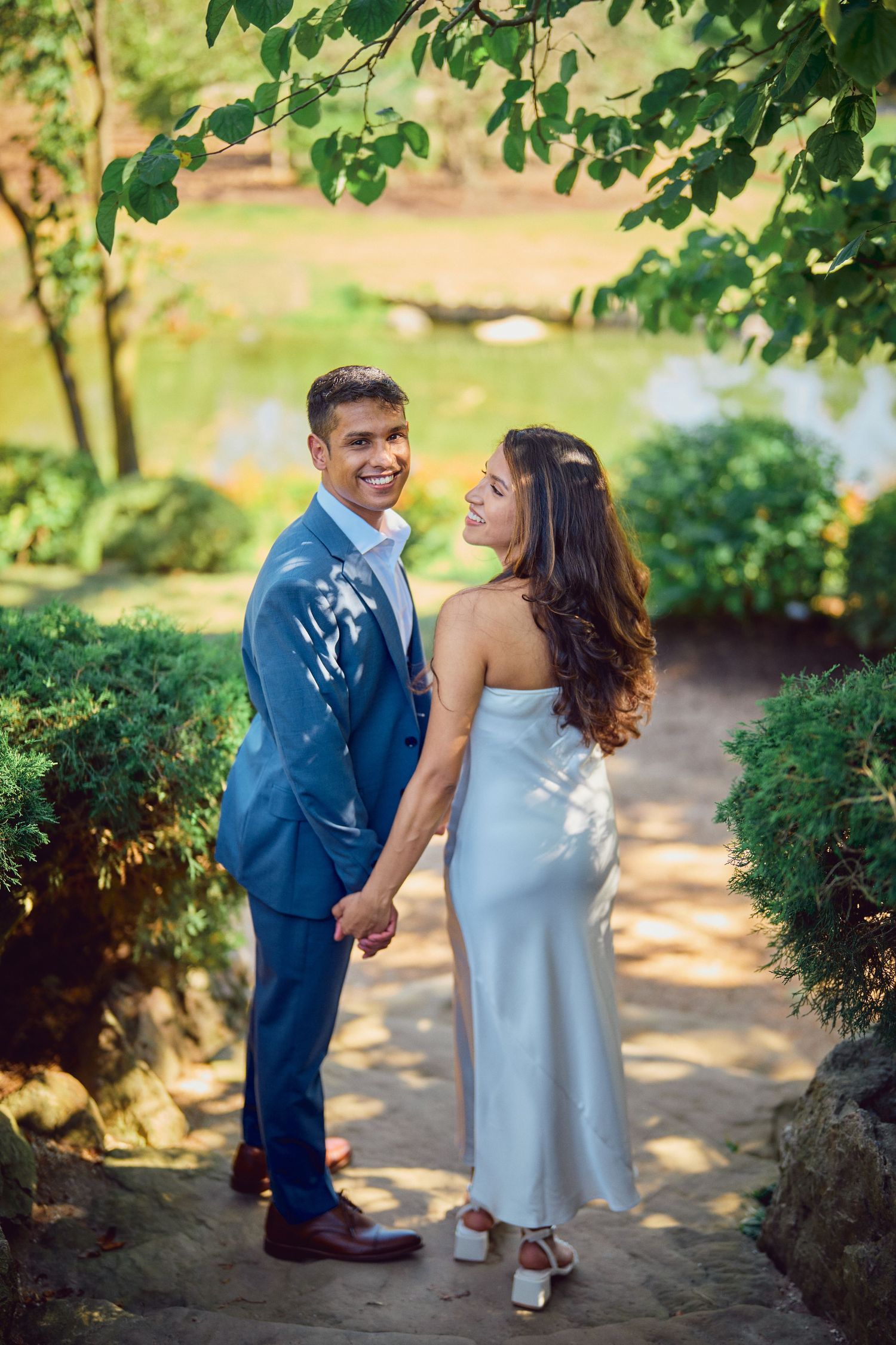 A couple holds hands on a garden path beneath dappled sunlight and green leaves on a summer day.