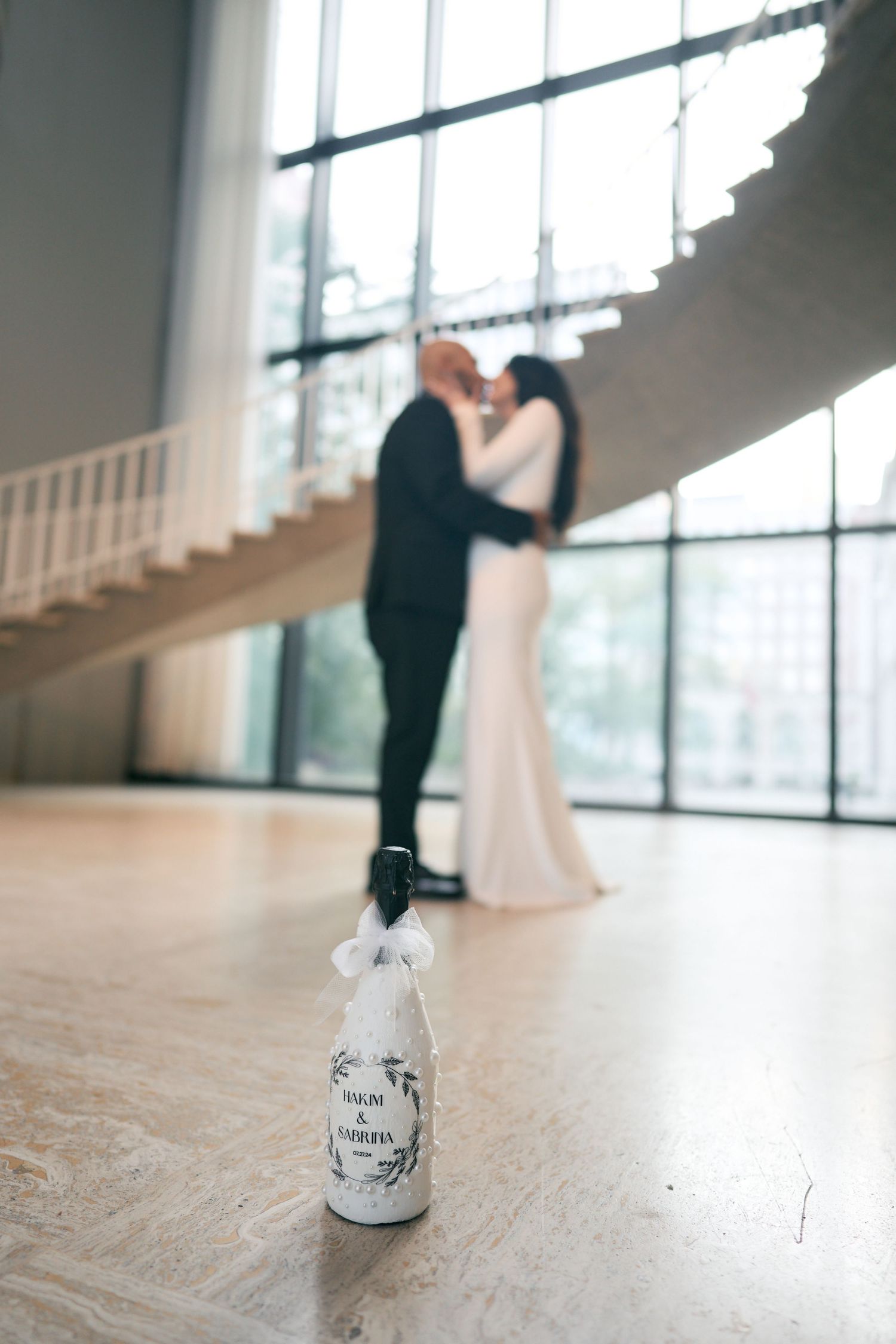 White decorative bottle sits in foreground while silhouettes of a couple embrace near tall windows in wedding venue.