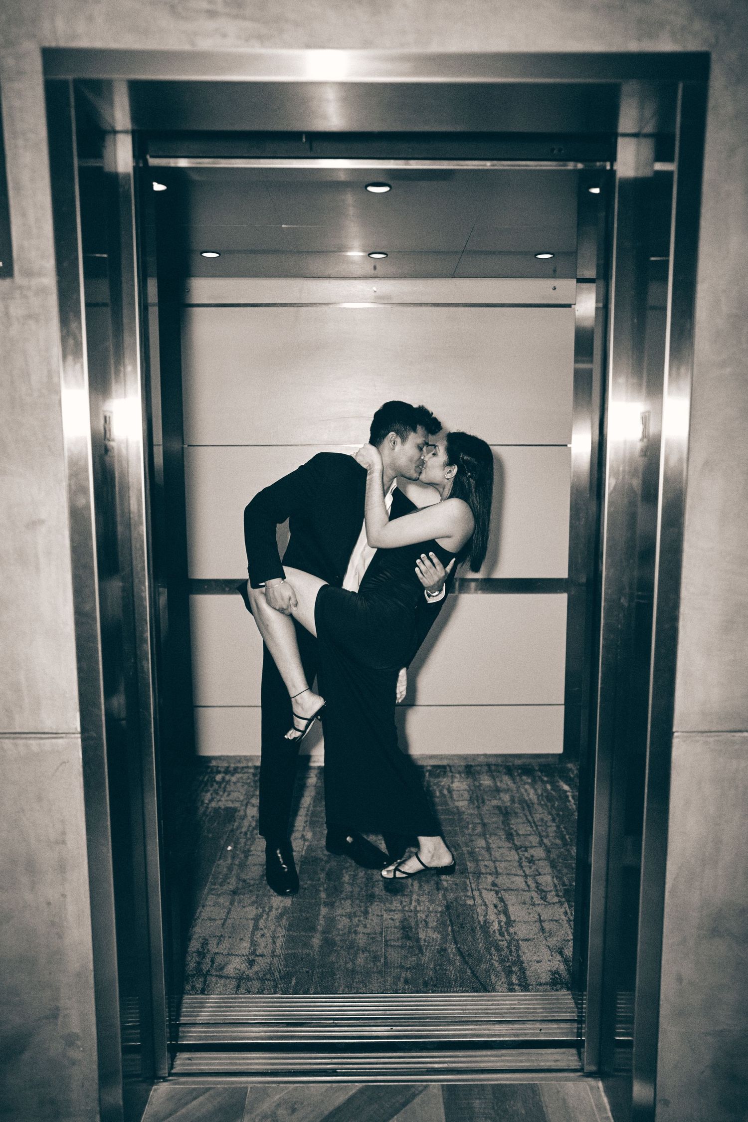 A romantic couple shares an intimate kiss in a modern elevator, captured in black and white photography.