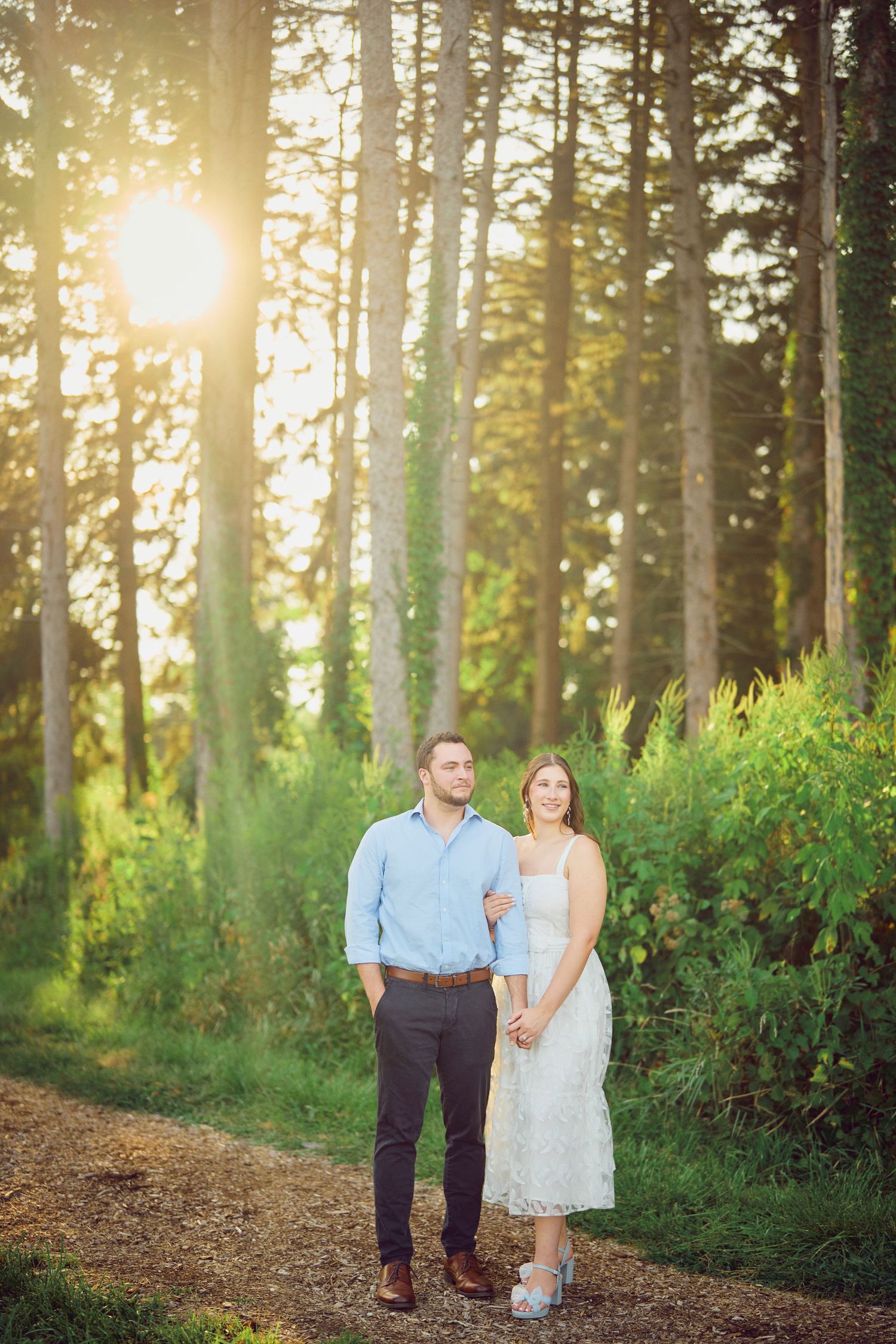A couple stands together on a path in a sunlit forest during golden hour.
