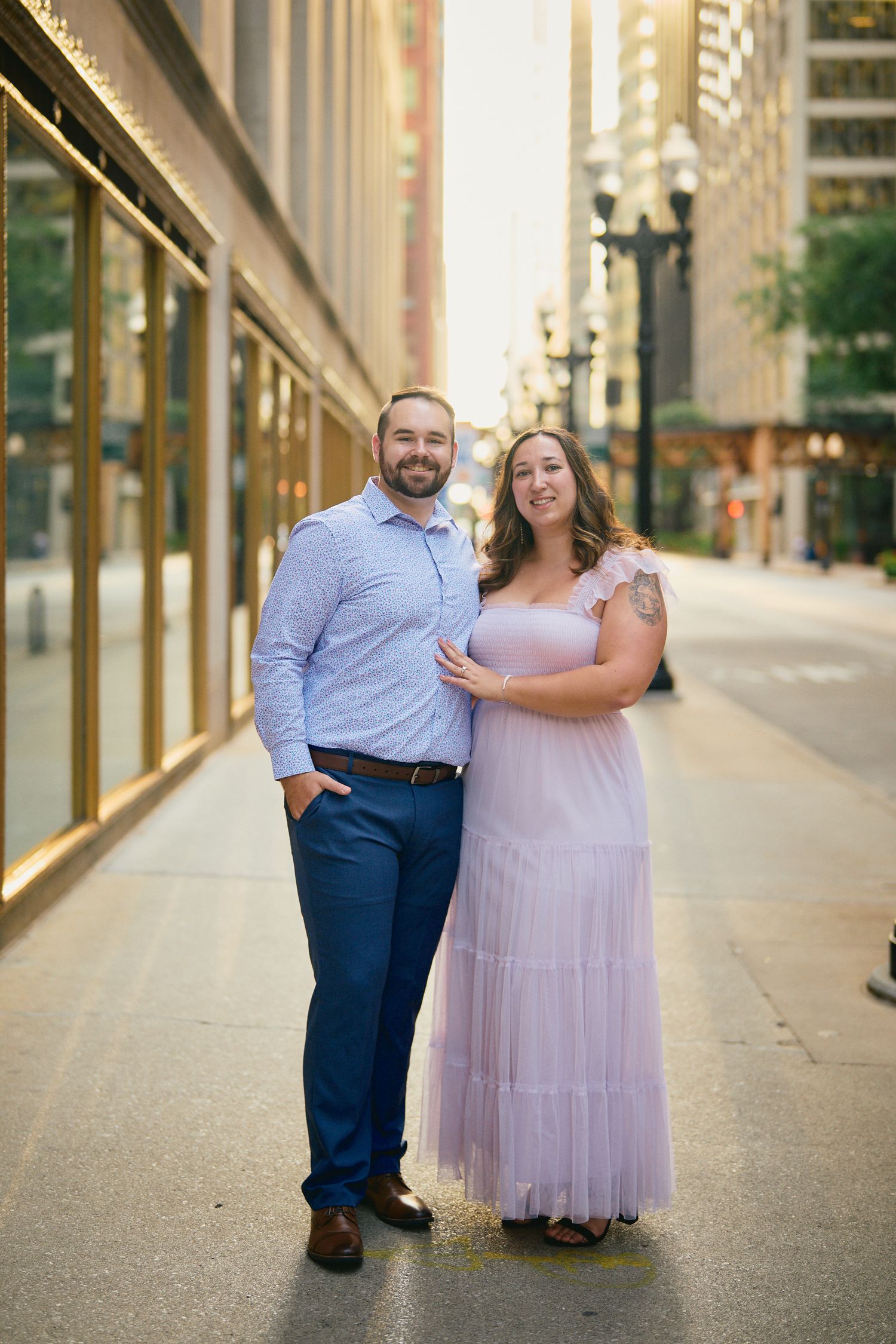A couple stands on a city sidewalk by decorative lampposts, with the woman wearing a flowing mauve dress.
