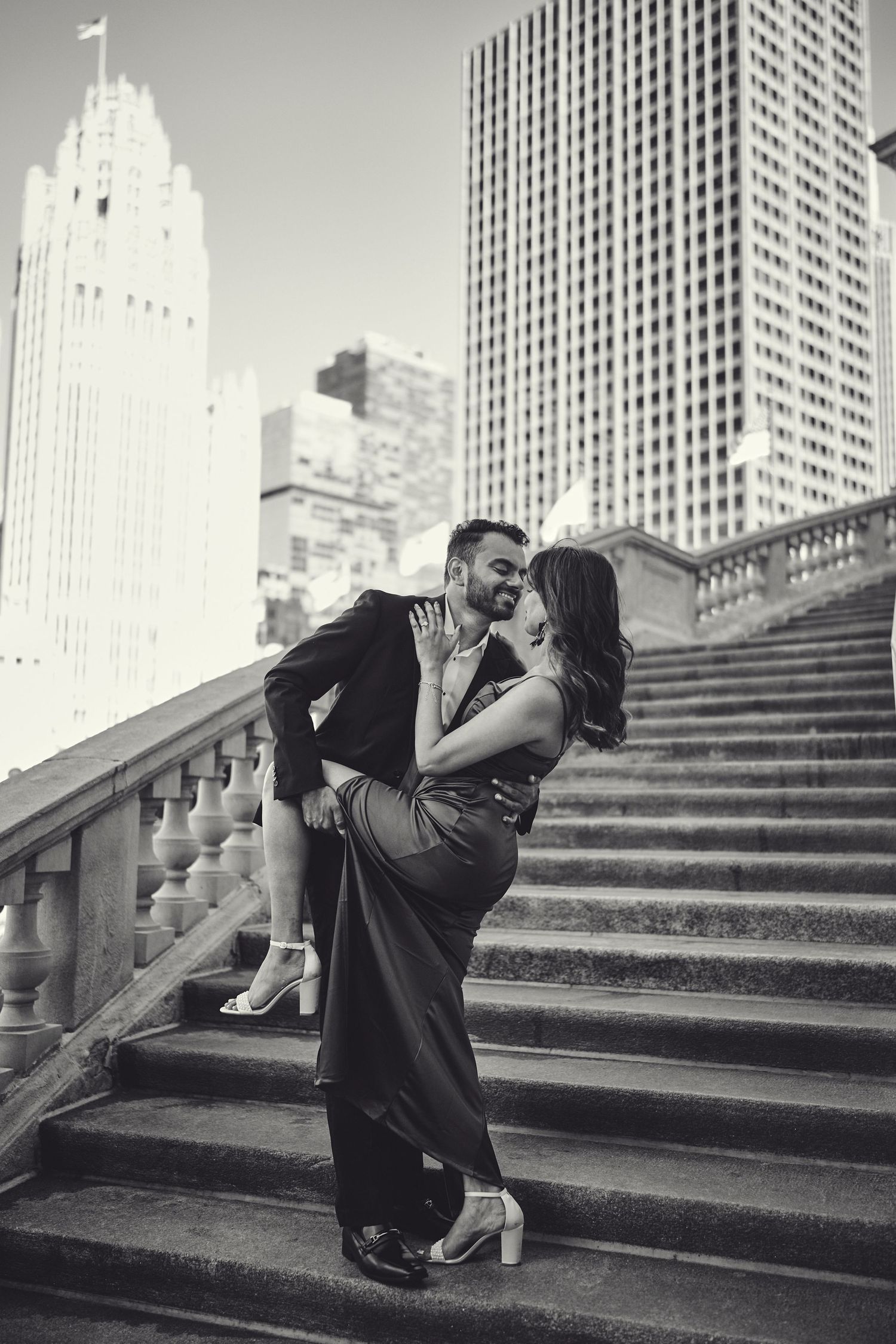 A romantic couple dancing on grand stone steps with tall city skyscrapers in the background in black and white.