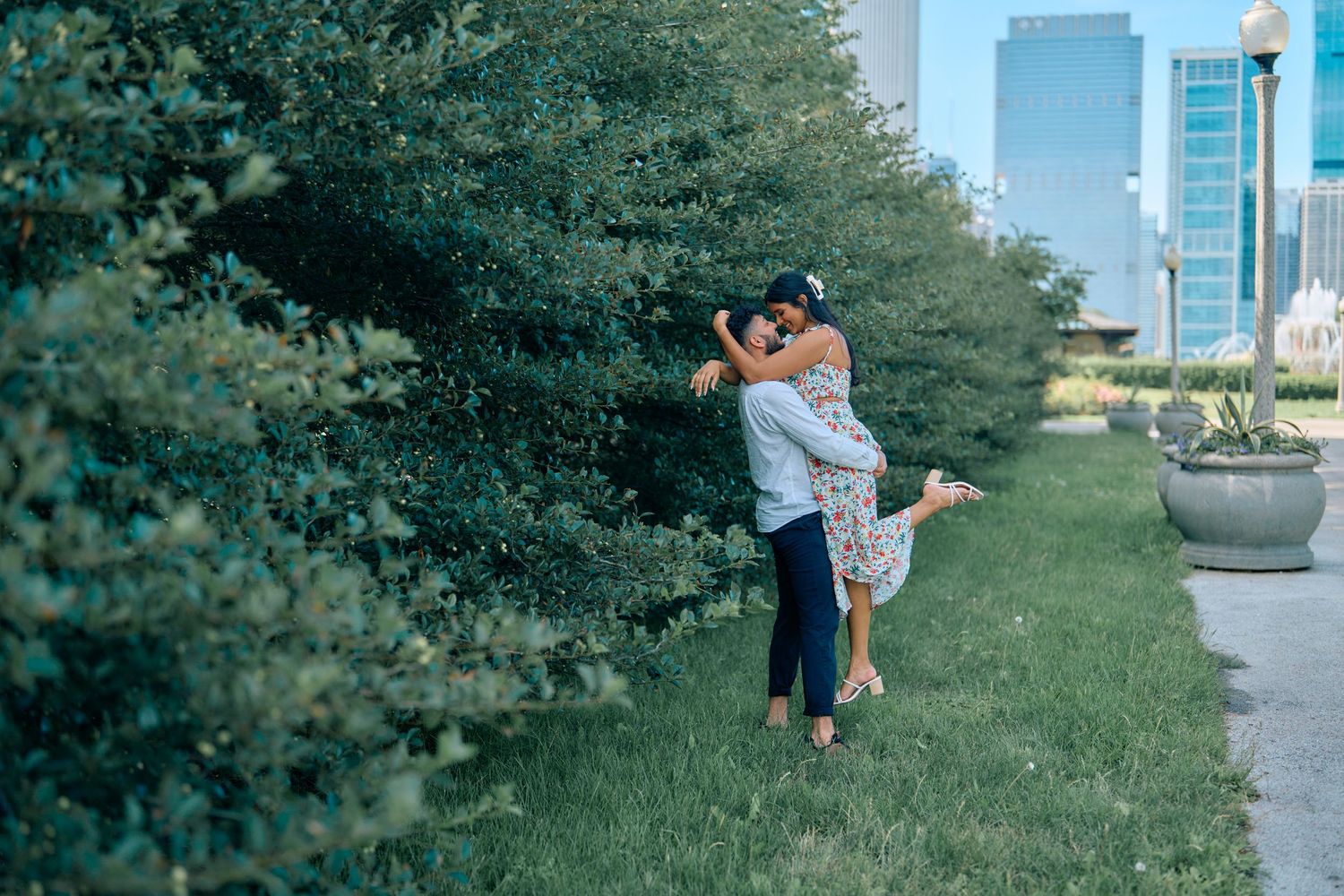 A couple embraces and shares a romantic moment in an urban park with city buildings visible in the background.