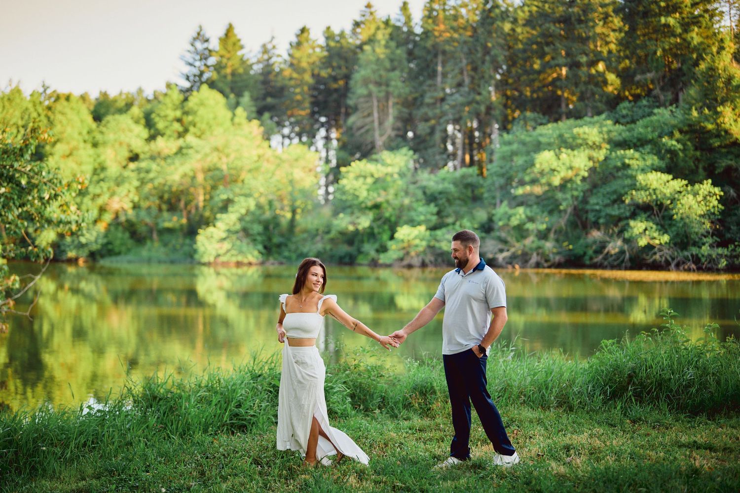 A romantic couple walks hand in hand by a serene lake surrounded by lush green forest on a sunny summer day.