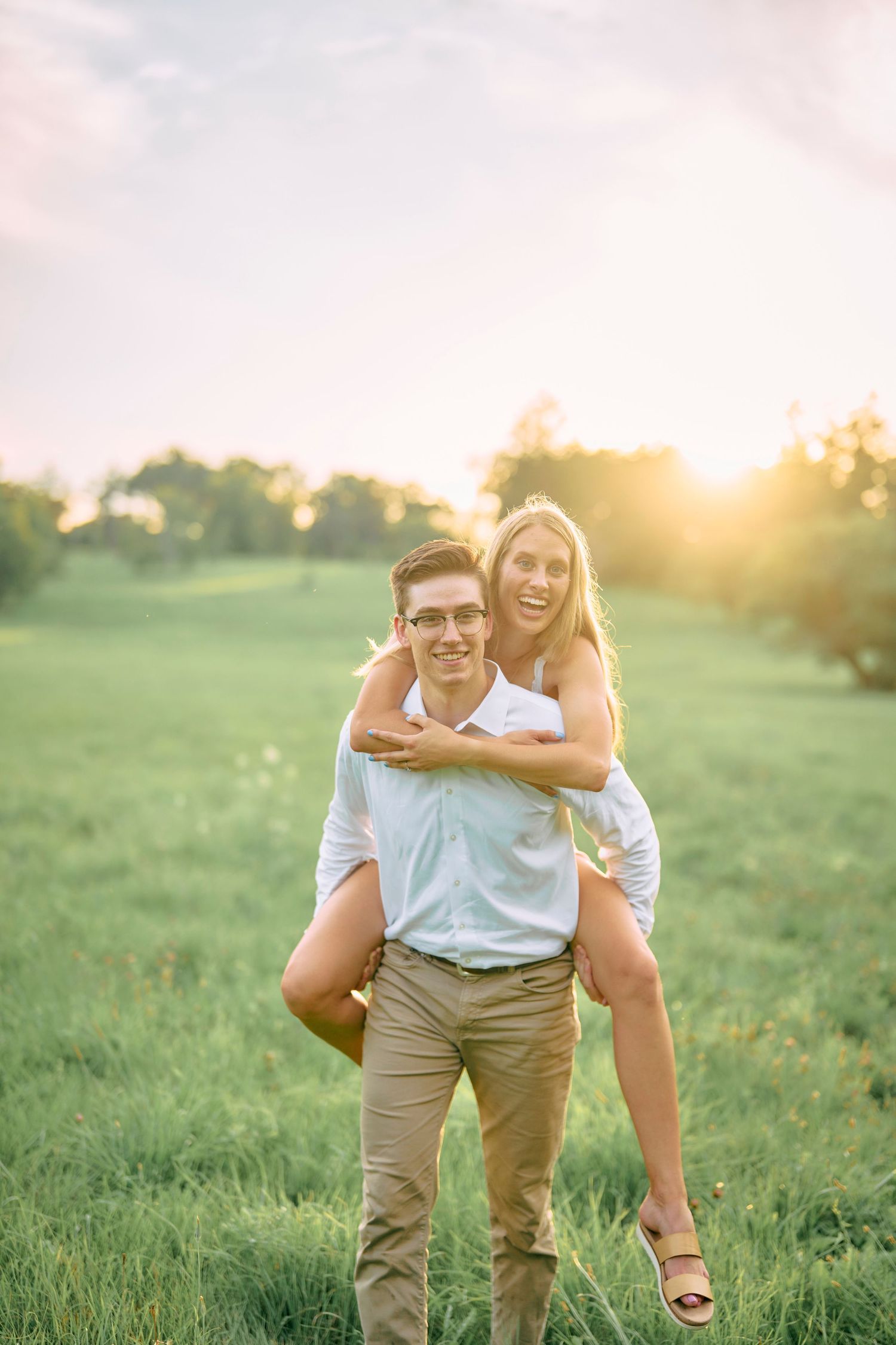 Couple shares a playful piggyback ride moment in a sunlit grassy field during golden hour.