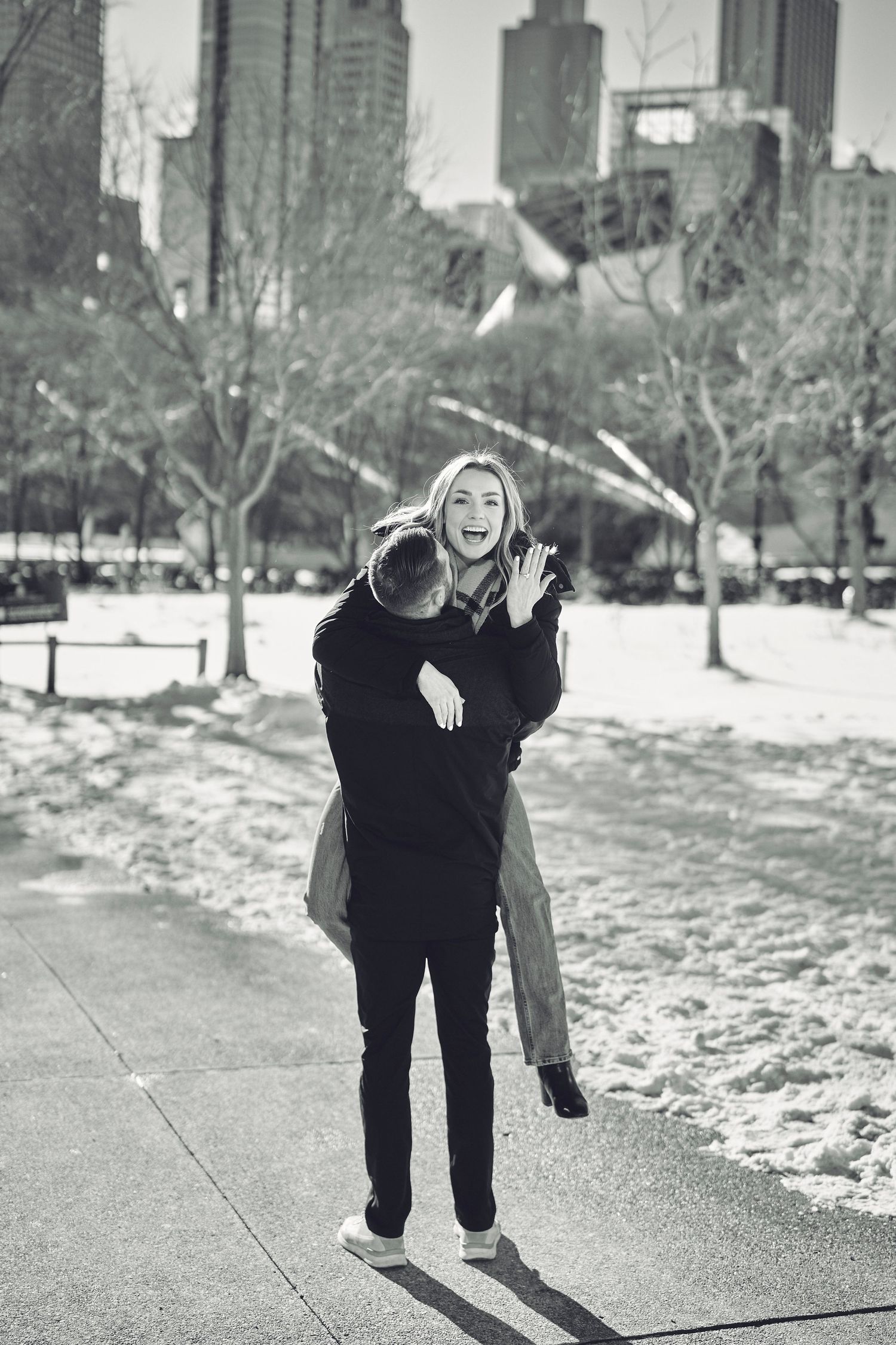 A joyful couple embraces on a snowy winter day in an urban park with bare trees and buildings in the background.