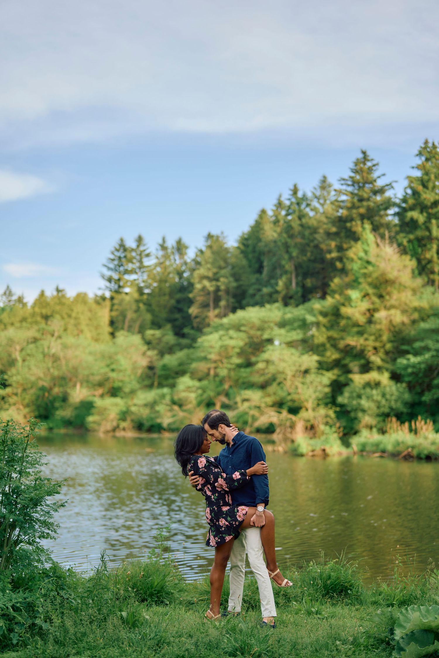 A couple embraces by a scenic lake surrounded by evergreen trees and lush greenery on a summer day.