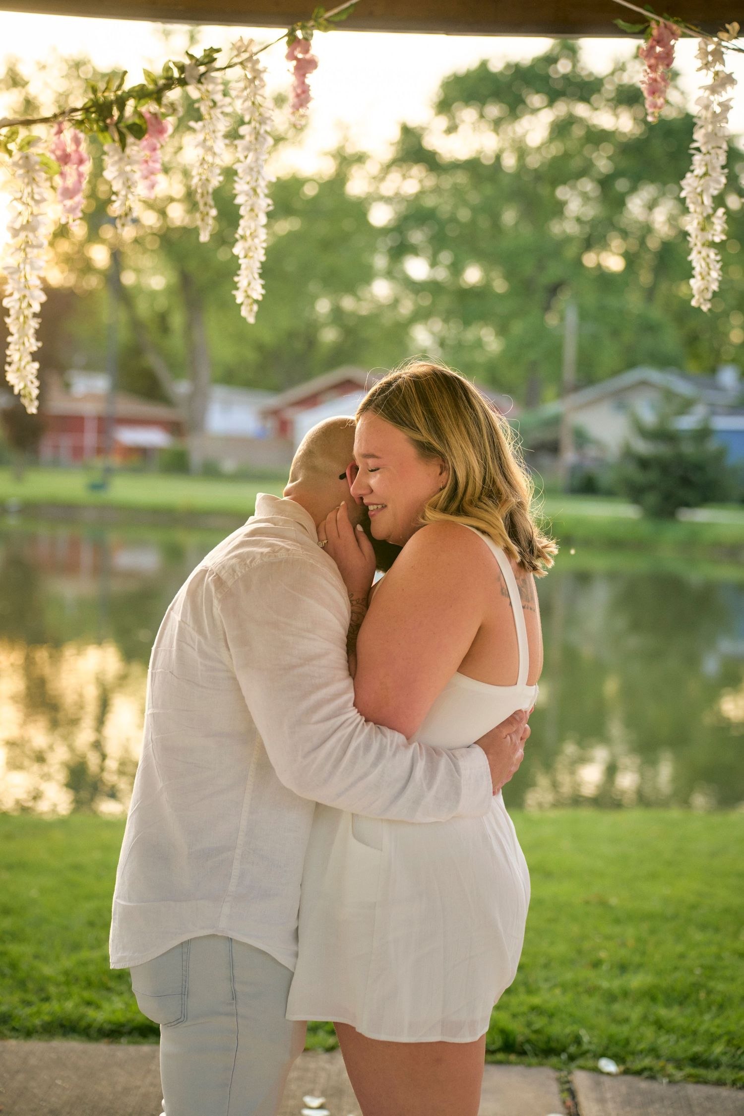A couple shares a tender moment at sunset by a lake under hanging wisteria flowers in a romantic outdoor setting.