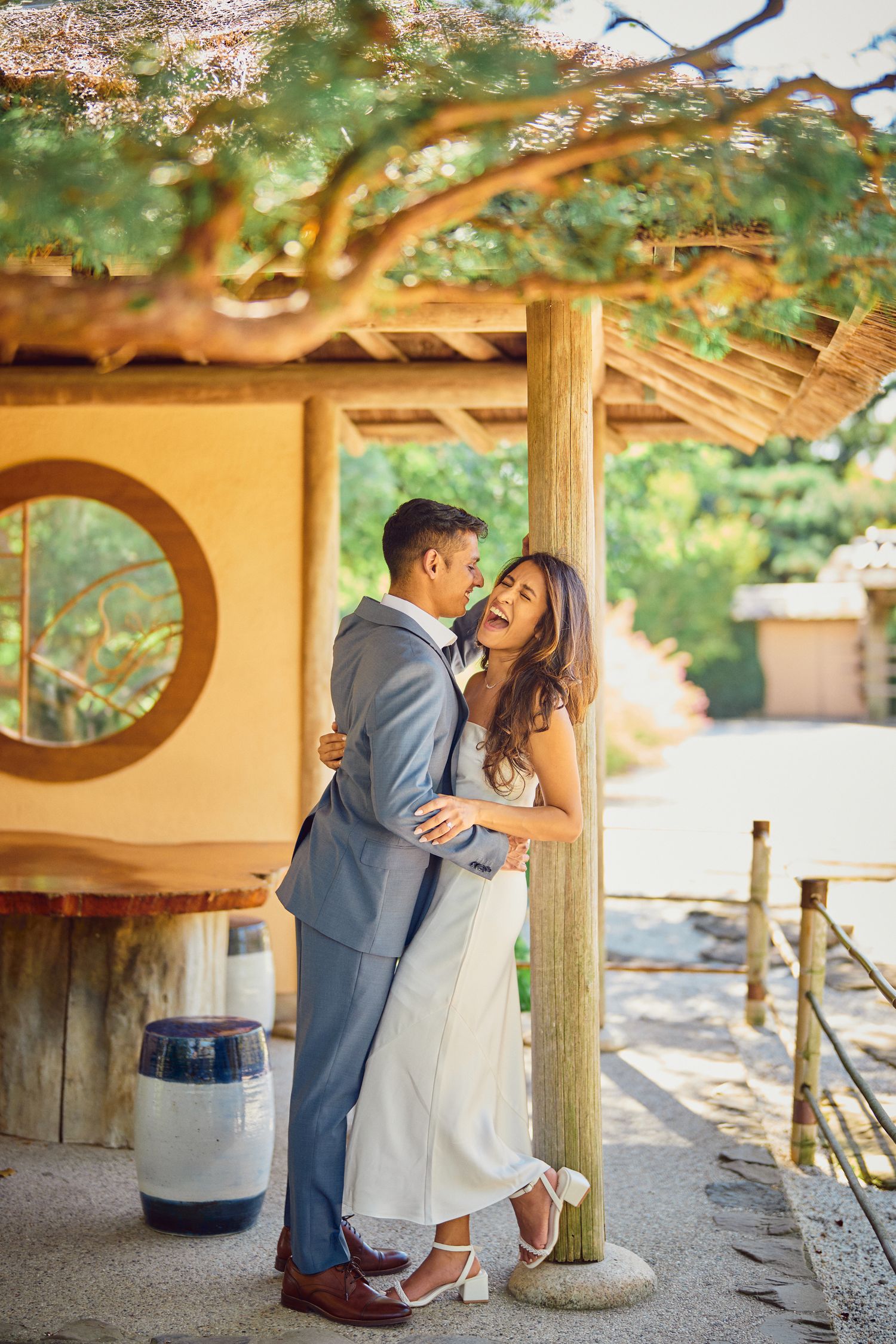 A couple share a romantic embrace under a wooden pergola at a rustic outdoor venue on a sunny day.