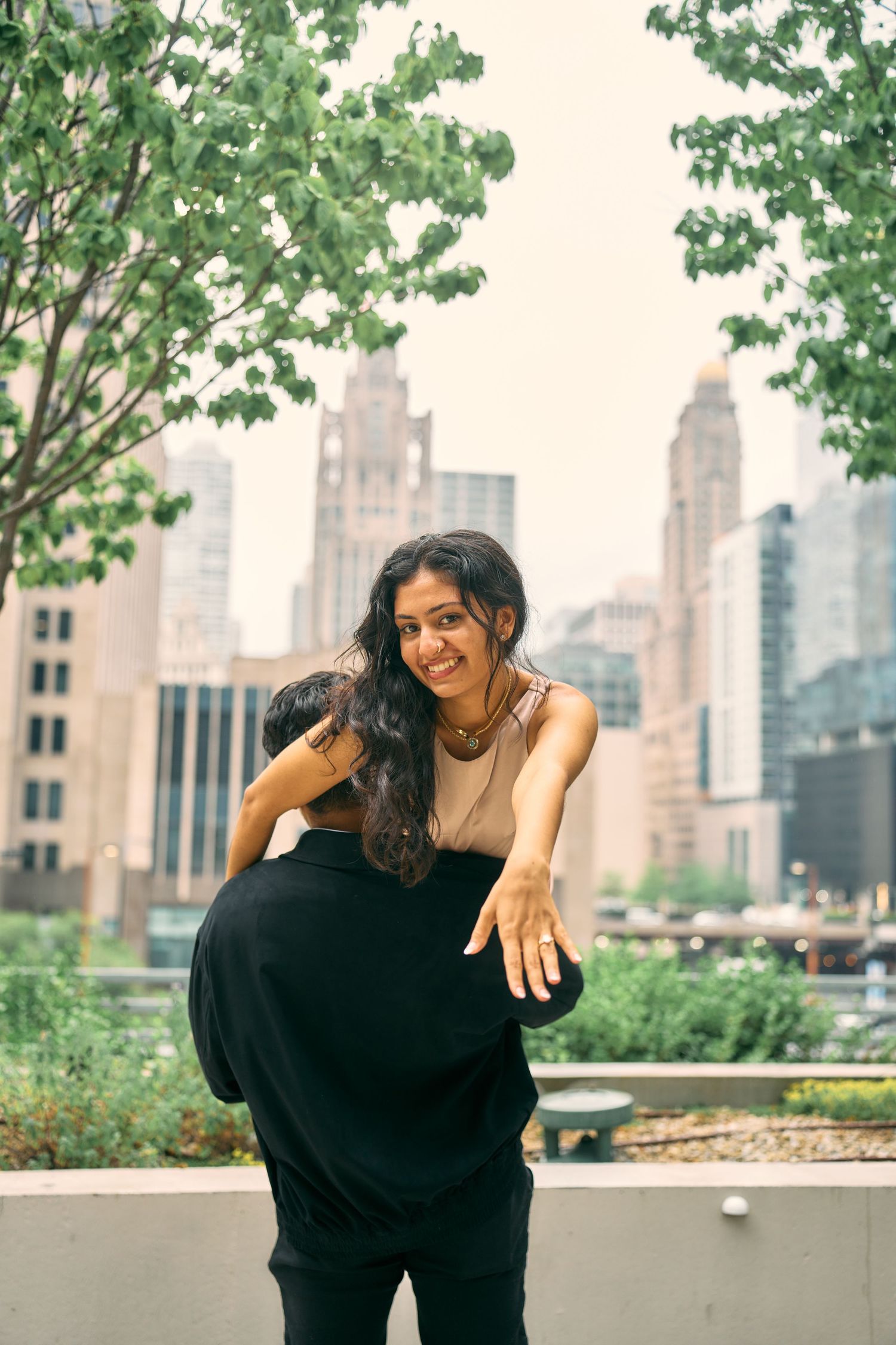 Person in black outfit smiles and poses against urban skyline with green trees in foreground and tall buildings in background.
