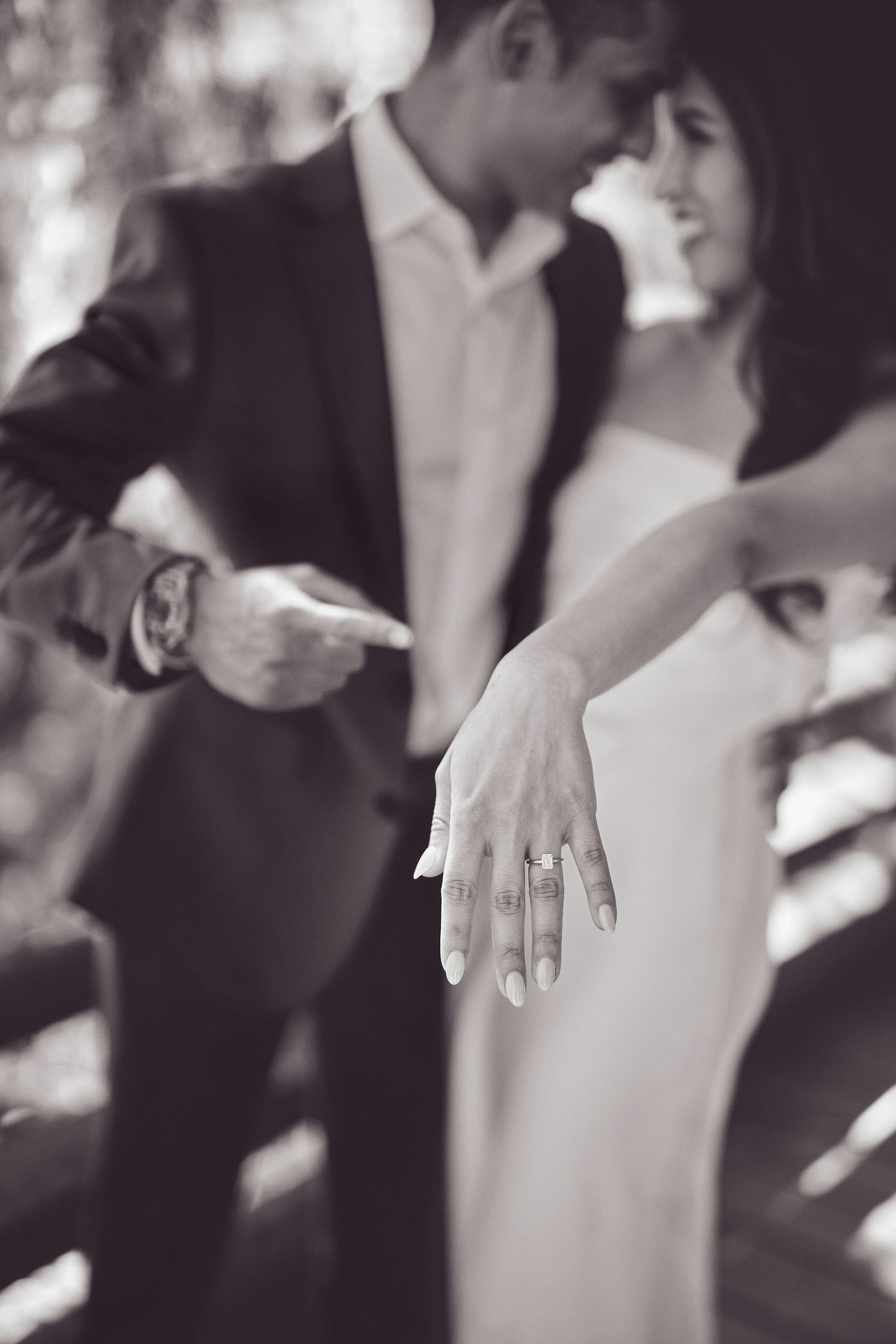 A romantic close-up photo in black and white showing a couple wearing formal attire with a sparkling engagement ring on display.