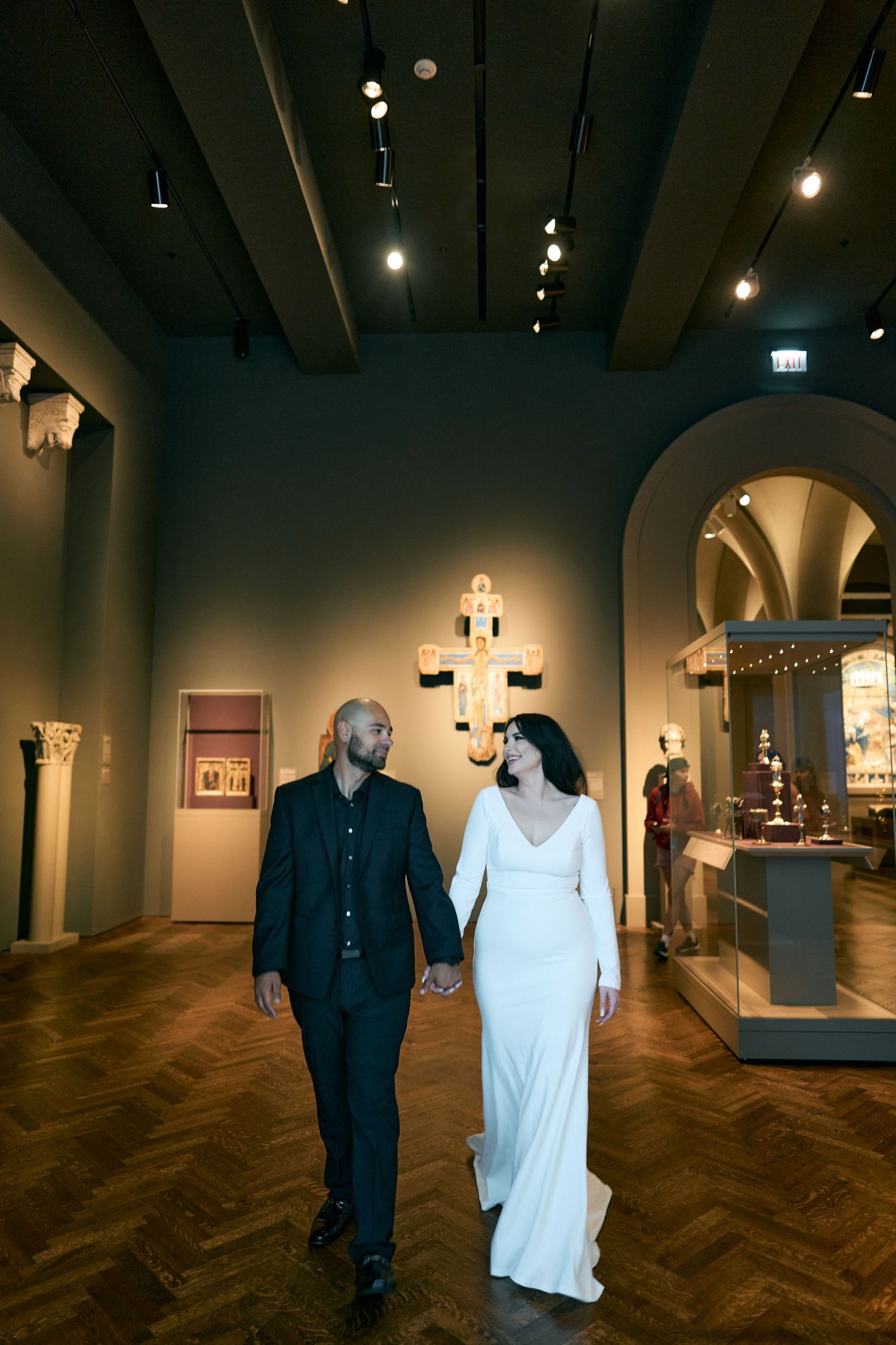 A couple walks hand in hand through an elegantly lit gallery space with a white gown and dark suit.