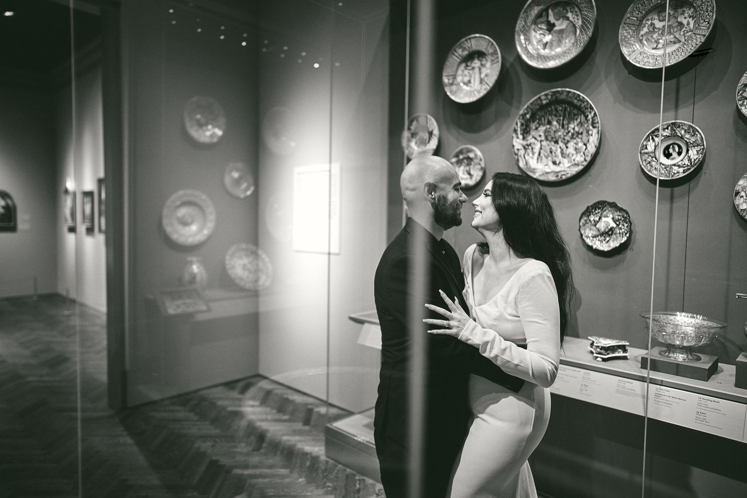 Two figures embrace in front of a gallery wall displaying decorative plates in a black and white photograph.
