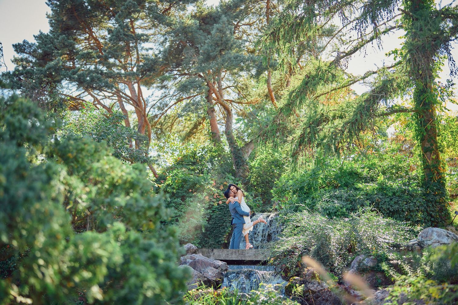 A couple shares an intimate moment while standing on a wooden platform surrounded by lush forest greenery.