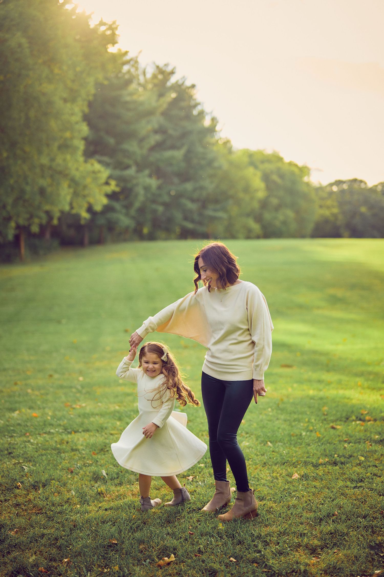 A parent and child dance together on a green lawn at sunset while wearing light colored clothes.