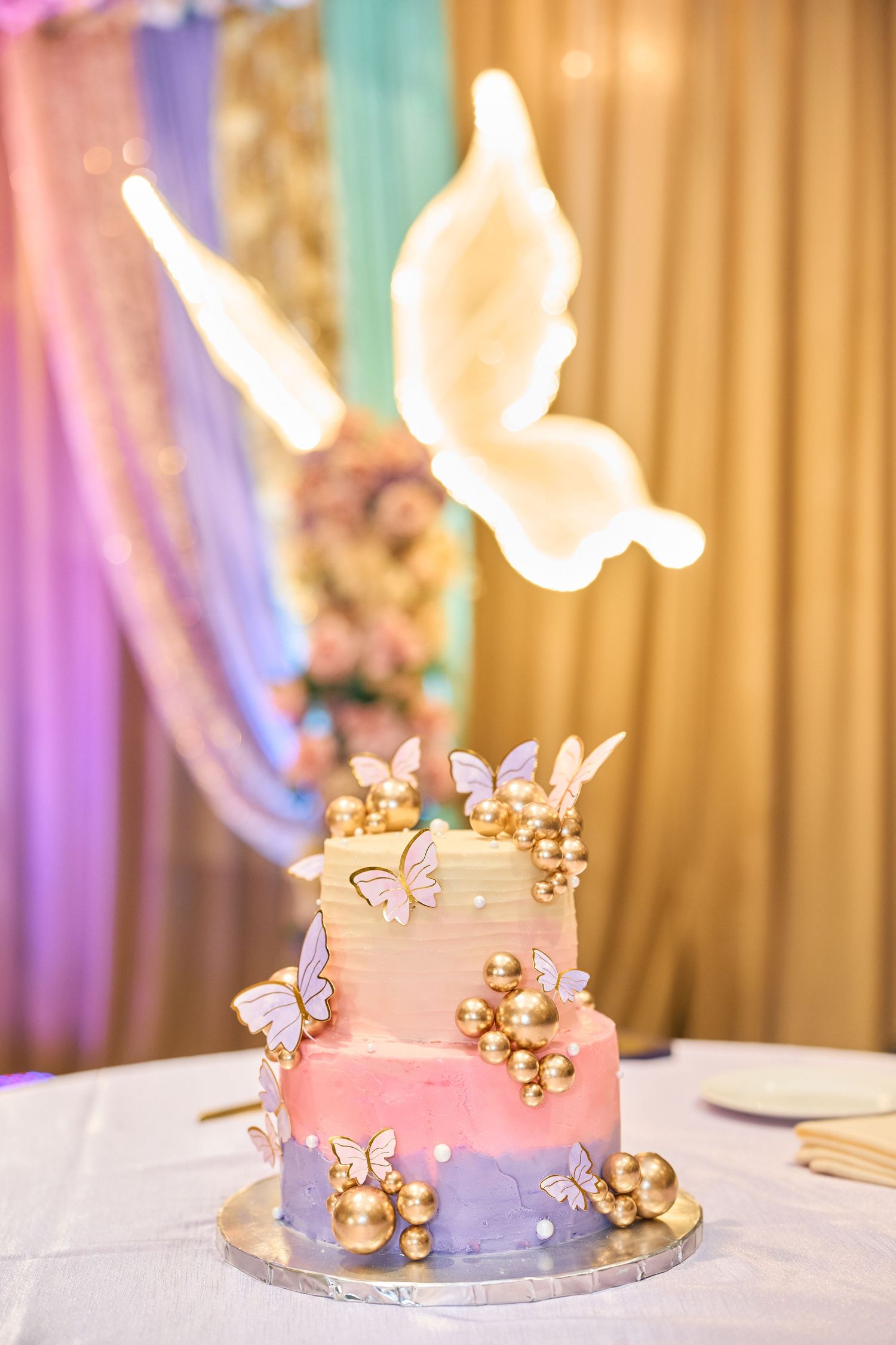 A tiered pink and purple ombre wedding cake decorated with gold spheres, butterflies and a large illuminated butterfly topper.