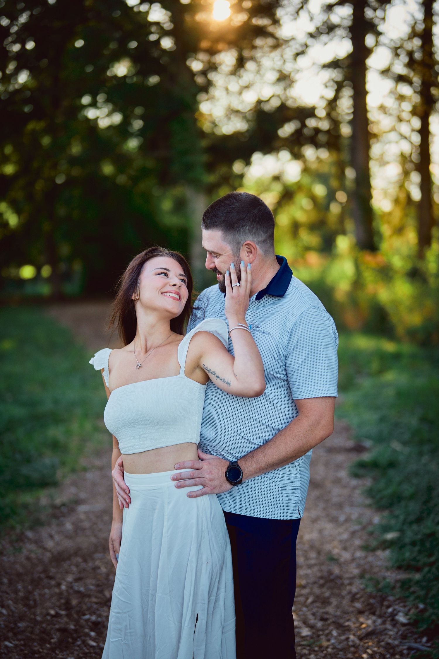 A romantic couple shares an intimate moment on a wooded path at sunset with golden light filtering through the trees.