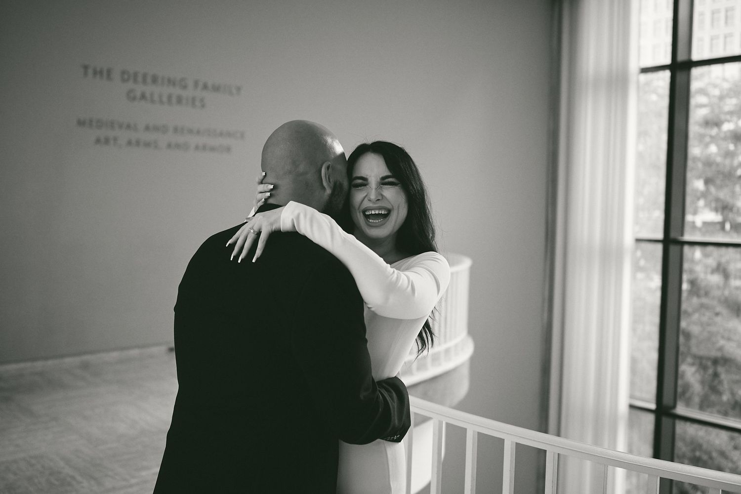 A couple embraces and laughs together in a bright minimalist room with white walls and a window.