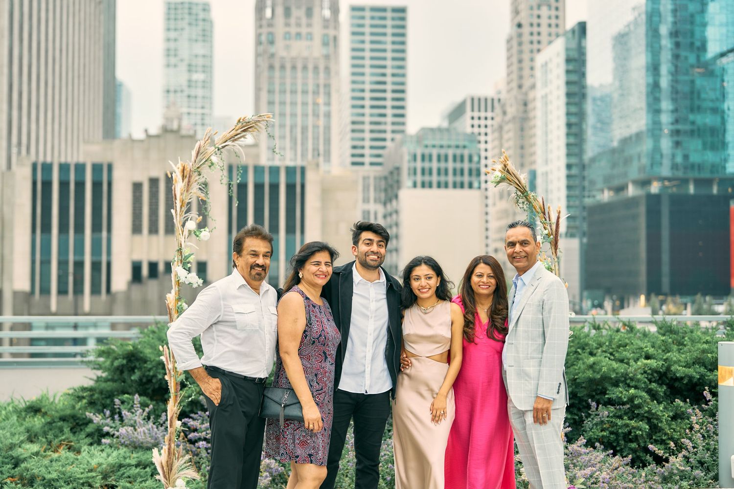 A group dressed in formal attire poses together in a lush rooftop garden with modern city skyscrapers in the background.