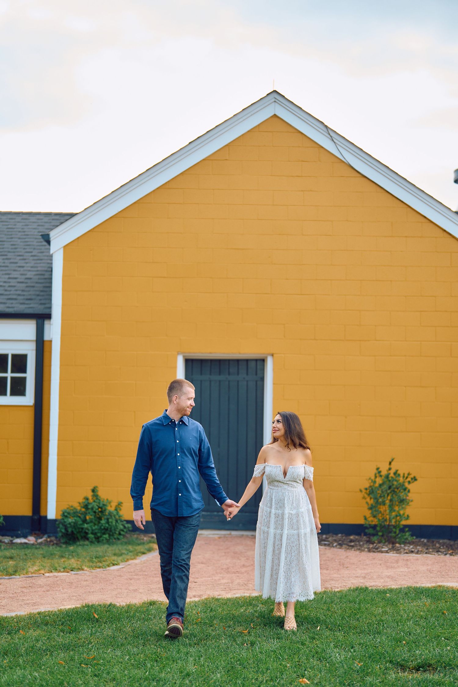 A couple holds hands while walking along a grassy path in front of a bright yellow building with a grey door.