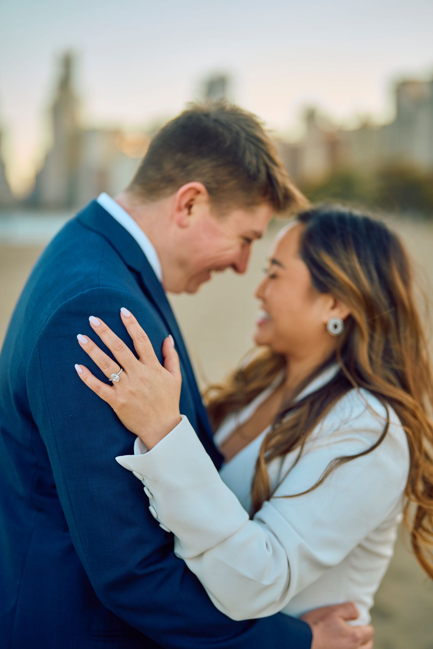 A newly engaged couple shares an intimate moment against a city skyline background while showing off the engagement ring.