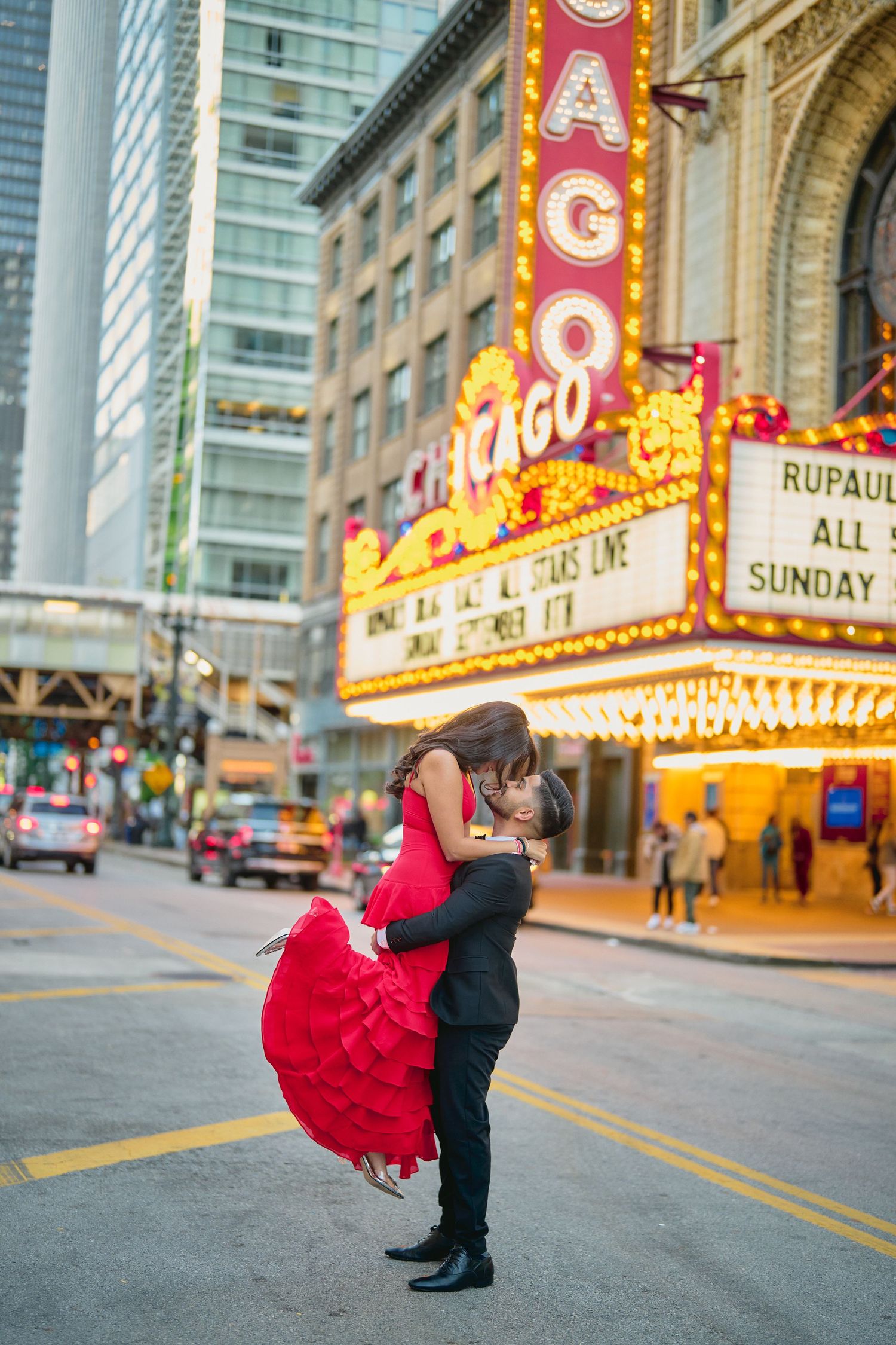 Couple embrace in front of illuminated Chicago Theatre marquee sign as woman's red dress flows dramatically.