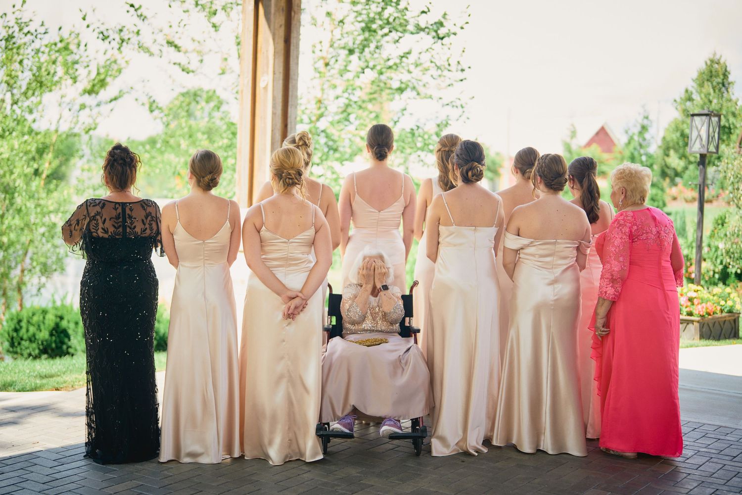 Group of people wearing formal dresses standing with their backs turned at outdoor venue in spring garden setting.