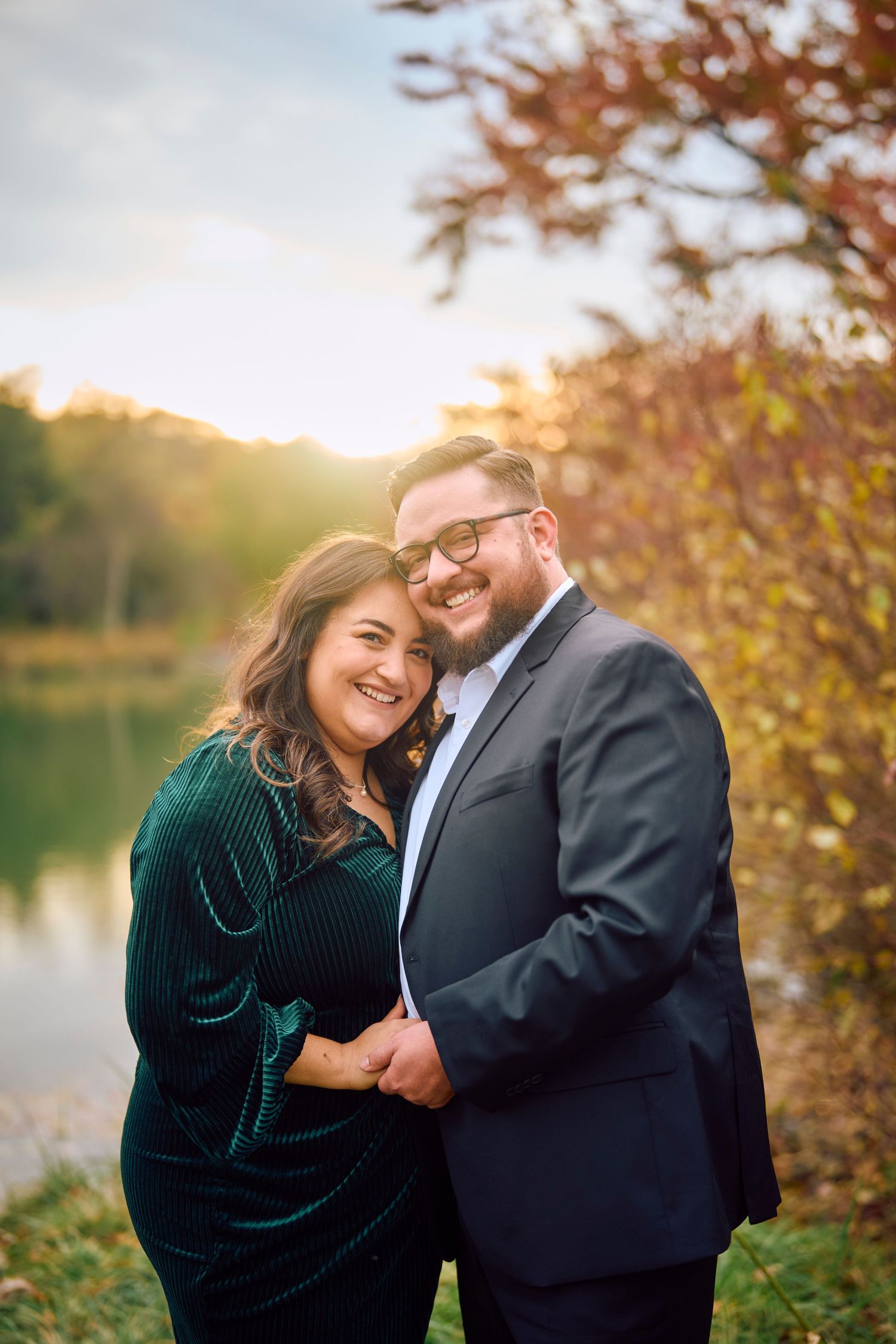 A couple in formal attire embraces by a lake during autumn sunset, with her in emerald velvet and him in a dark suit.