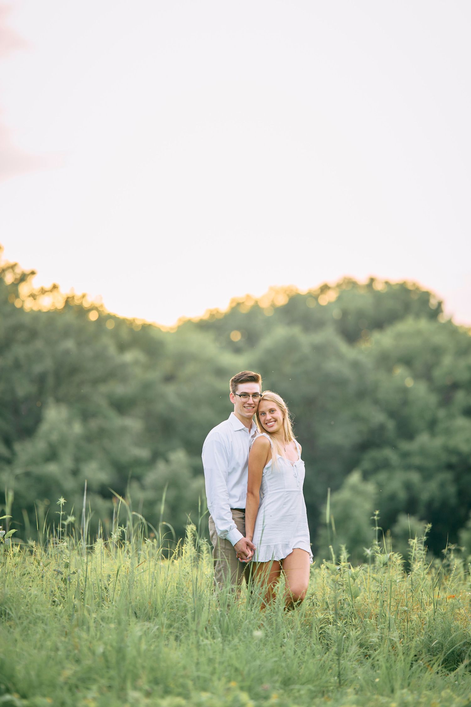 A couple in white outfits stands together in a summer meadow at sunset with trees in the background.