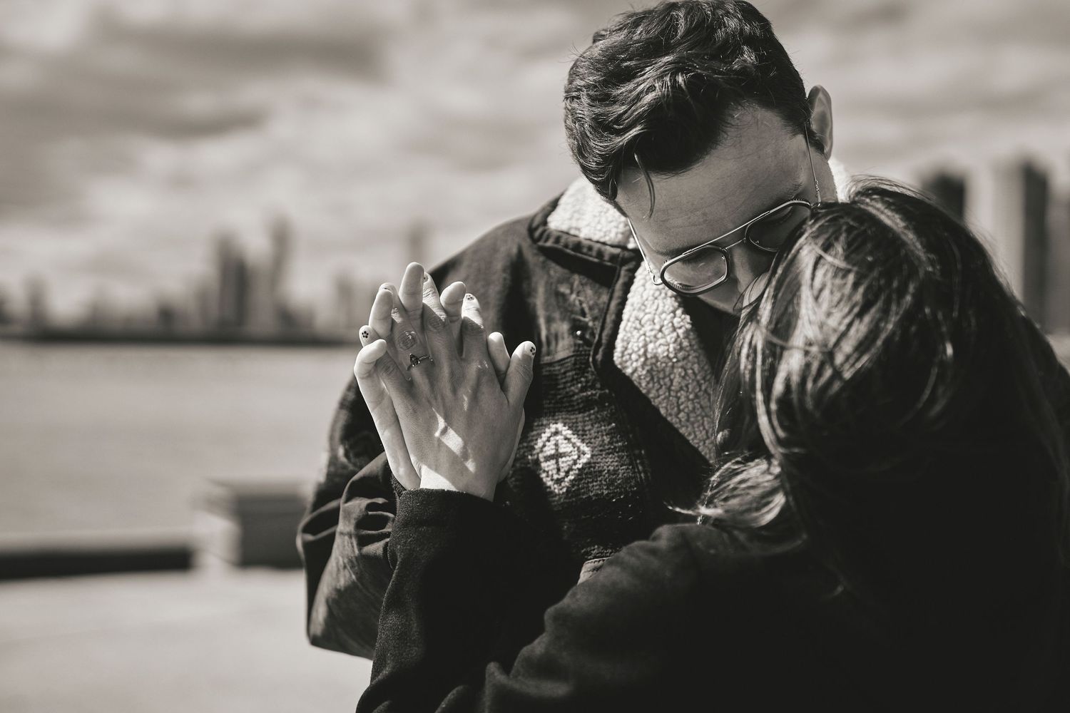 A person in a leather jacket lights a cigarette against an urban waterfront backdrop in black and white.
