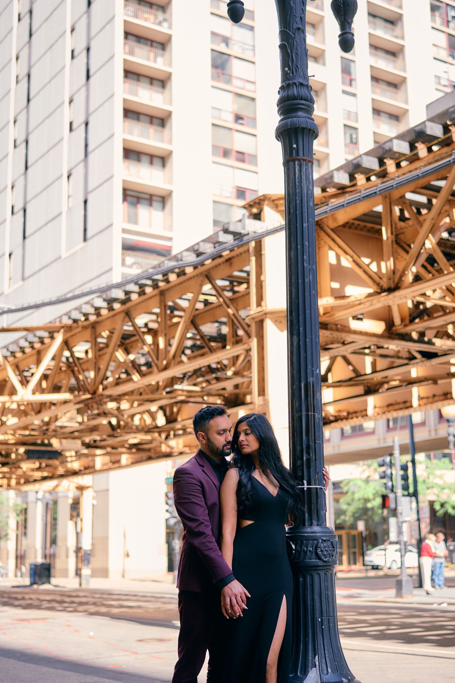 A couple poses romantically against a vintage lamp post beneath elevated train tracks in an urban setting.