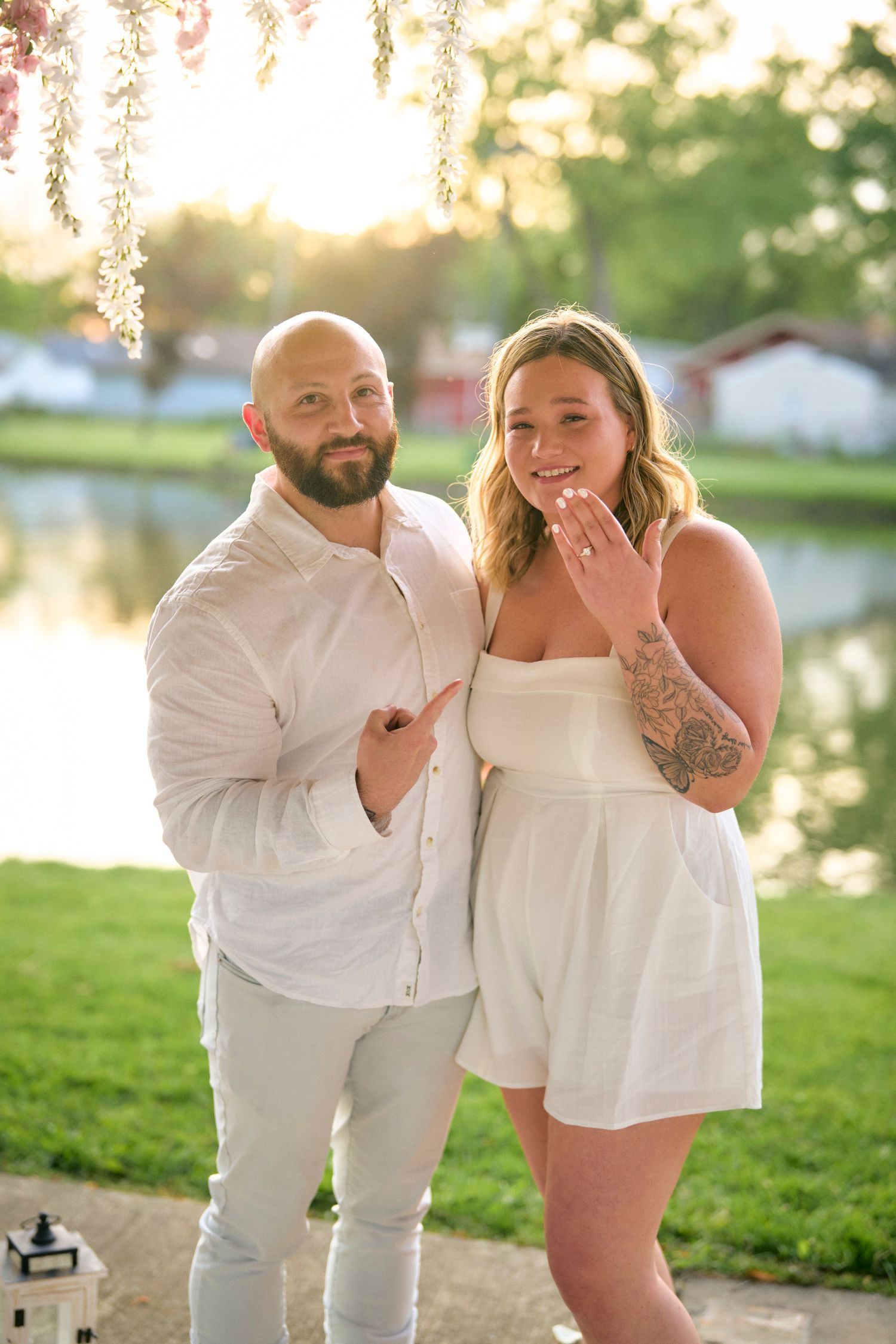 Couple in white outfits posing outdoors at sunset near a lake with spring blossoms overhead.