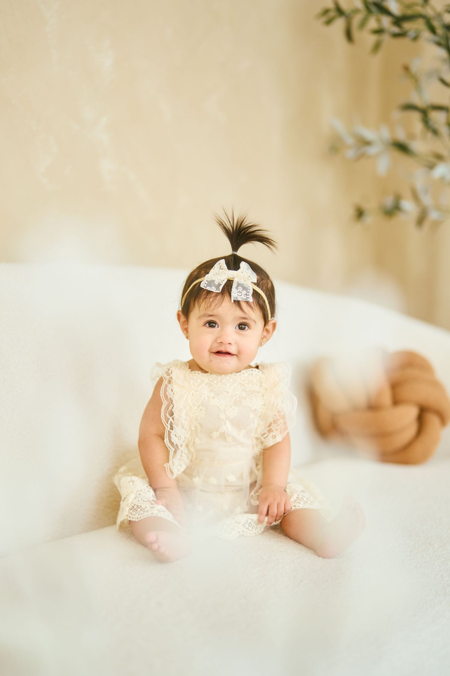 A baby wearing a cream lace dress and headband sits on a white fluffy rug in a studio setting with soft lighting.